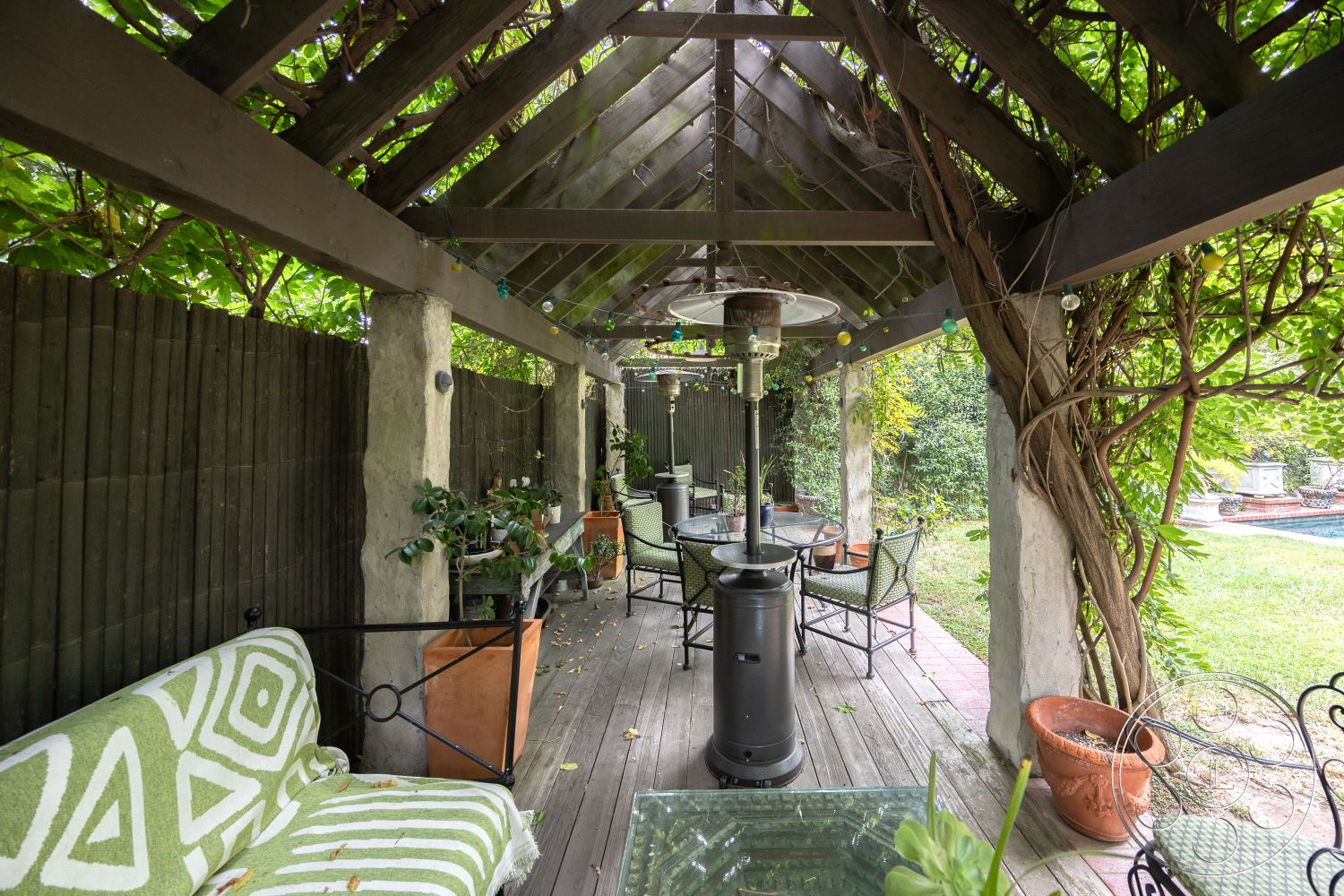Mansion 2 - potted plants, outdoor seating area, landscape view, concrete wall, relaxation area, landscaped backyard, open-air environment, garden setting, outdoor heater, textured plaster wall, urban outdoor retreat, wooden pergola structure, exterior patio space, decking wood floor, metal outdoor table, green patterned outdoor cushions, hanging vine plants, string lights for ambiance