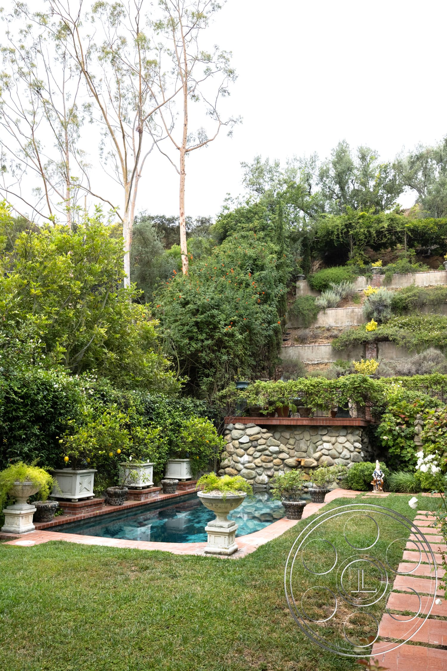 Mansion 2 - natural stone texture, exterior garden setting, suburban backyard retreat, rectangular pool with concrete border, stone feature wall with brick cap, grass lawn area, decorative planters with flowering plants, surrounding tall trees with leafy foliage, bricks used for patio walkway, lush garden landscape with varied greenery, pool area with clear water, elevated hillside background, interior/exterior transition area, elegant stone masonry details, modern outdoor living space