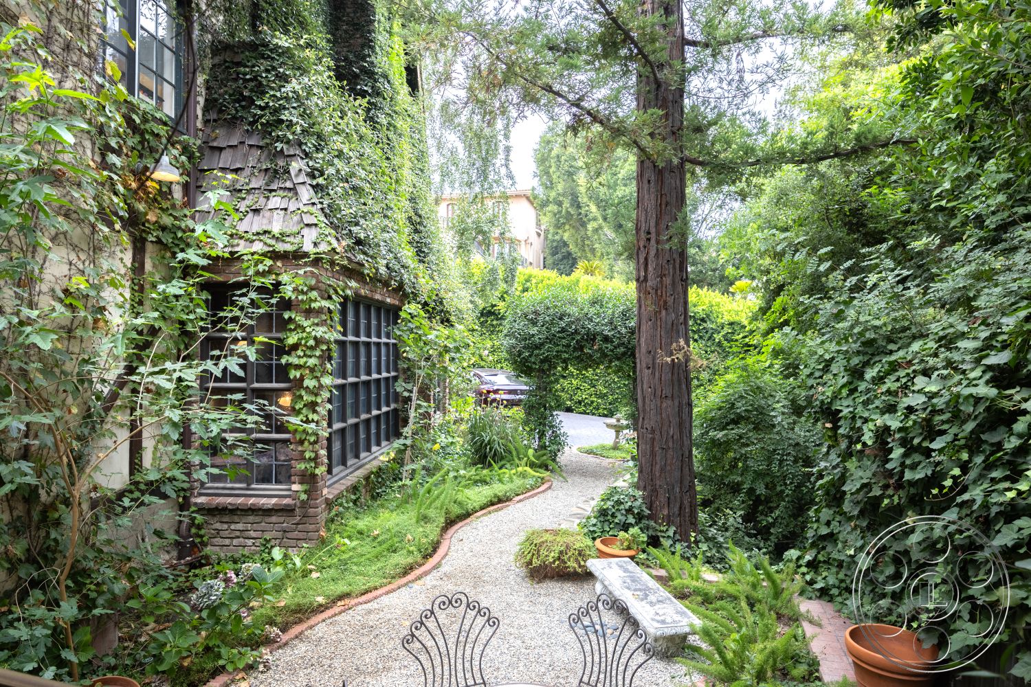 Mansion 2 - exterior, outdoor seating area, natural wood beams, surrounded by lush greenery, large pane windows, native landscaping, stone wall cladding, wood shingle roof, shrub-lined pathway, circular gravel path, vertical wooden planters, overgrown ivy facade, decorative wrought iron bench, concrete garden bench, rustic cottage style, intimate garden space
