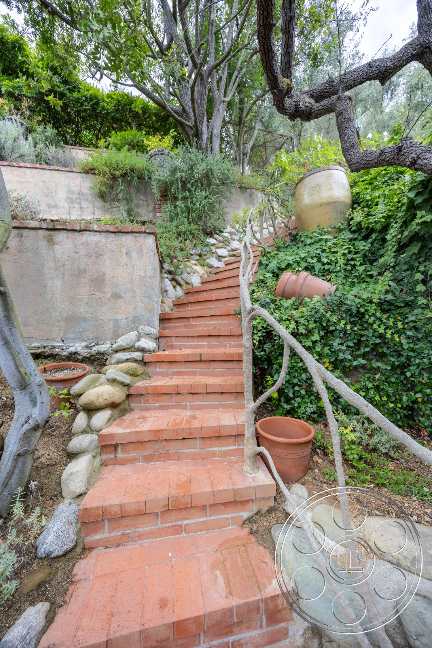 Mansion 2 - exterior, garden setting, hillside location, lush greenery backdrop, wooden handrail, organic shapes in landscaping, terraced garden design, outdoor brick stairs, red clay paver steps, natural stone pathway border, exposed earth surrounding, intricate garden landscaping, rustic clay flower pots, textured earthen wall, curved stair geometry