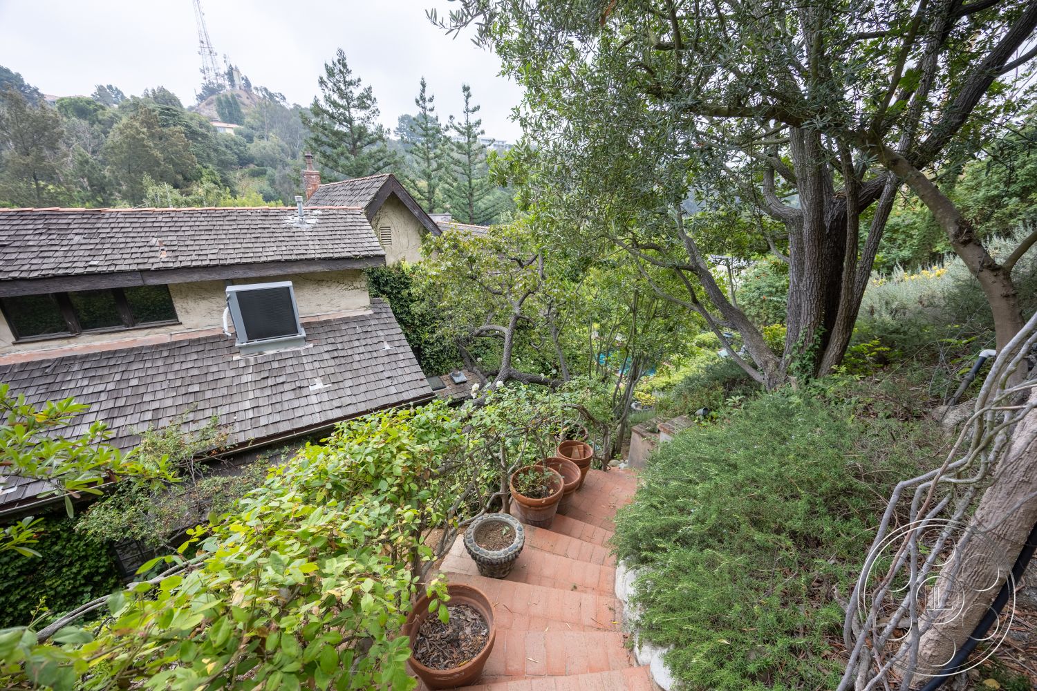 Mansion 2 - exterior, natural stone accents, lush greenery, mature trees, wooden window frames, slate roof, drought-resistant landscaping, mountain view setting, stucco wall finish, exposed brick chimney, curved garden pathway, sloped hillside location, wooden deck railing, red clay tile steps, stone garden retaining wall, terracotta plant pots, rustic outdoor ambiance