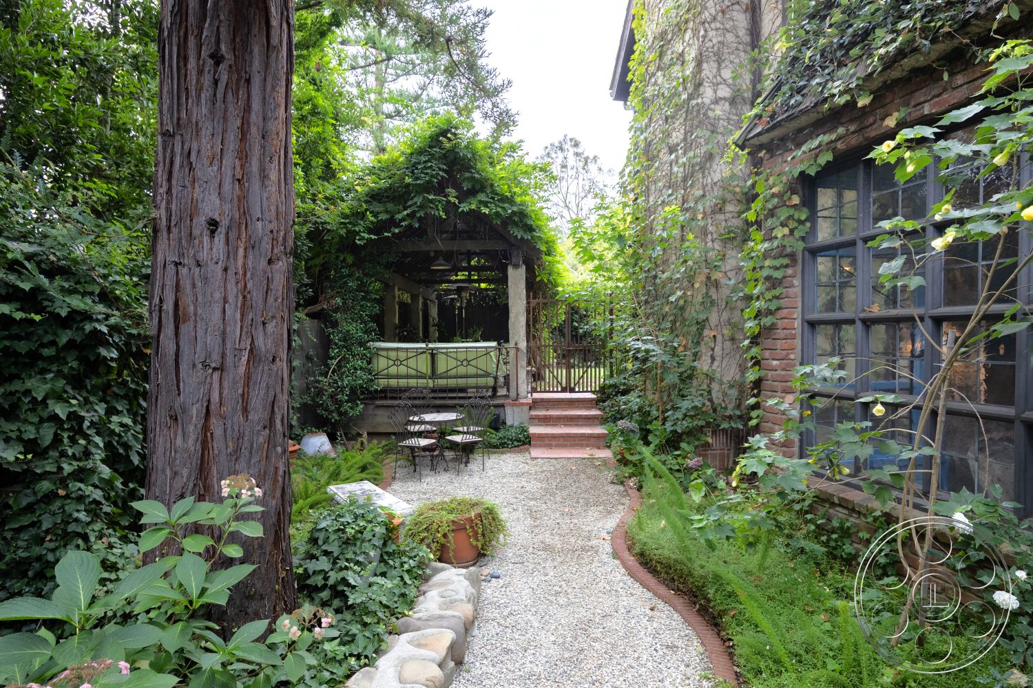Mansion 2 - exterior, exposed wood beams, ivy-covered walls, soft green foliage, wrought iron gate, private outdoor retreat, lush garden landscaping, flowering shrub borders, cobblestone patio area, stone garden pathway, natural stone wall treatment, brick house exterior, wooden trellis structure, gravel walkways, small outdoor seating area, rustic farmhouse style, secluded garden setting, curved flower bed, overhanging trees