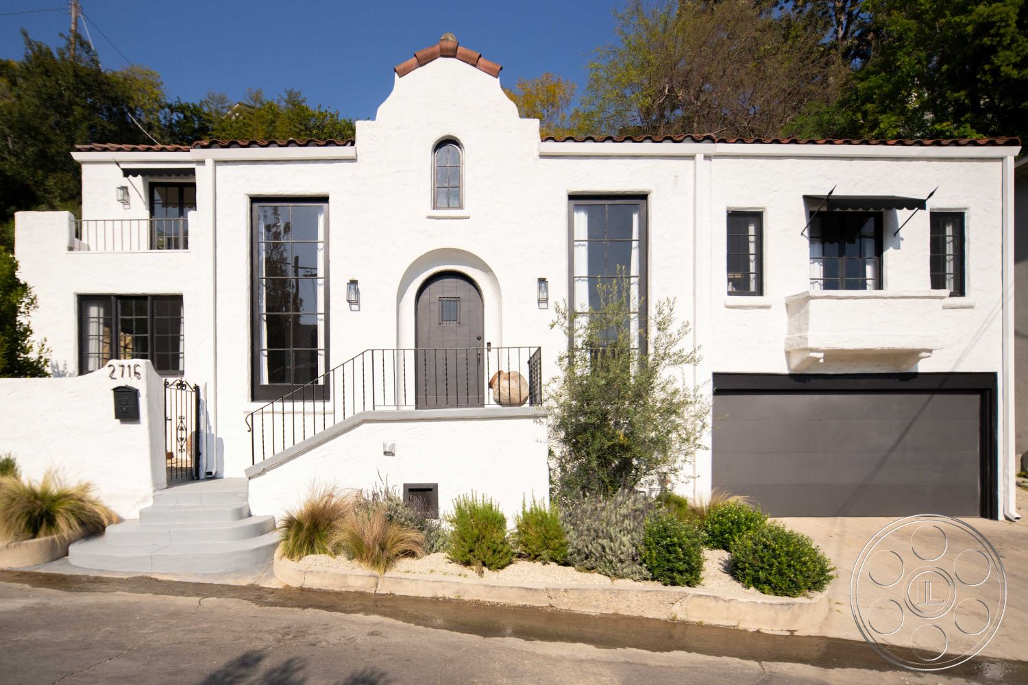 Spanish 166 - 2 car garage, paved driveway, symmetrical facade, Mediterranean architectural style, wrought iron railing, exterior stucco walls, sloped driveway, landscaped garden beds, urban hillside setting, minimalist outdoor lighting, concrete porch steps, round arched doorway, black glass exterior windows, flat roof with terracotta tile accents, horizontal wooden garage door, Spanish architecture, curbside