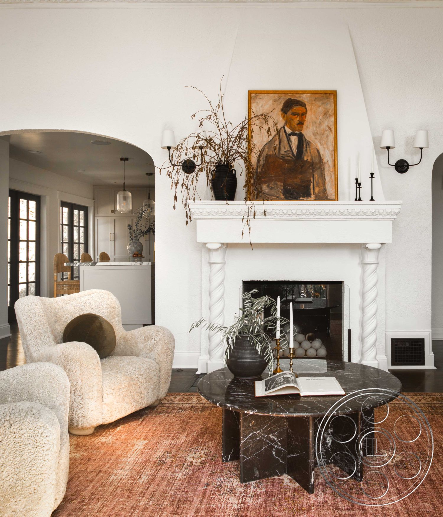 Spanish 166 - columns, textured area rug, dark hardwood flooring, open-concept living space, minimalist decor elements, white textured plaster walls, interior living room, natural light through large windows, traditional home design, marble fireplace mantel, exposed black marble coffee table, curved upholstered armchair, traditional white crown molding, artwork featuring a portrait, decorative vase with dried plants, candlesticks on fireplace mantel, arched doorways leading to kitchen, black wall sconces, decorative wooden accents in kitchen