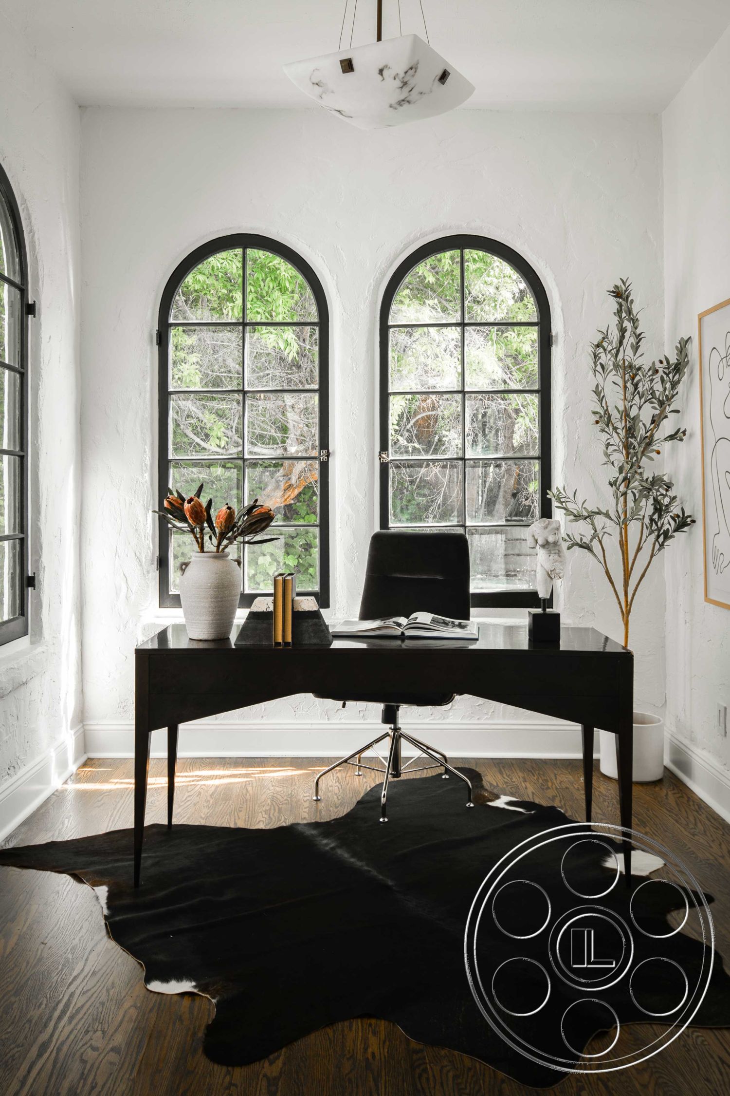 Spanish 166 - neutral color palette, hardwood flooring, artwork on walls, contemporary design elements, white textured plaster walls, natural light-filled workspace, minimalist office decor, black cowhide area rug, arched windows with black frames, marble pendant light, modern black desk with tapered legs, greenery outside windows, chrome office chair