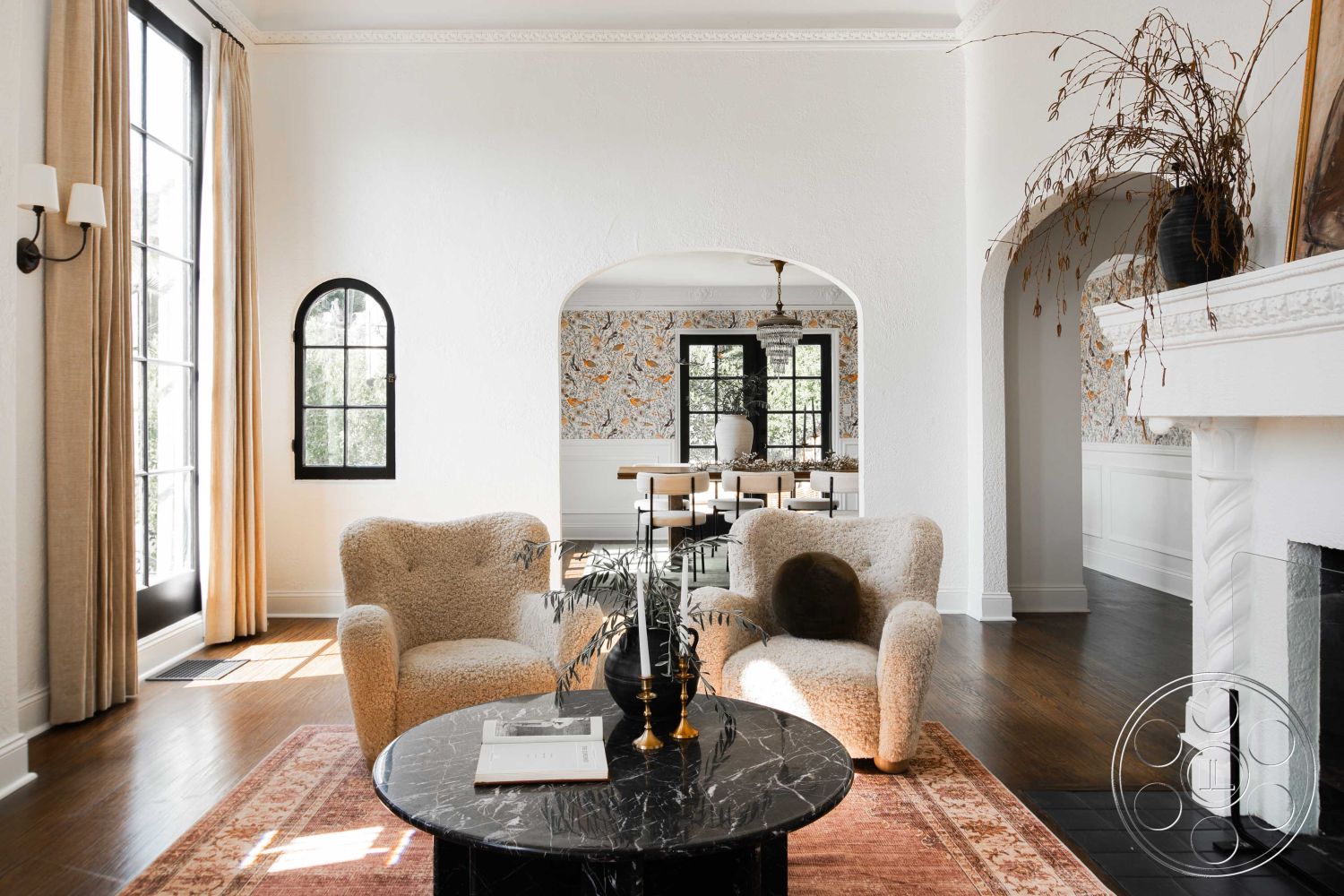 Spanish 166 - fireplace, columns, arched doorways, textured area rug, crown molding details, chandelier light fixture, hardwood flooring living room, exposed white plaster walls, black steel window frames, dark marble coffee table, beige boucle armchairs, cream drapery curtains, wall art with floral patterns, dining area with wooden table, black ceramic vase decor