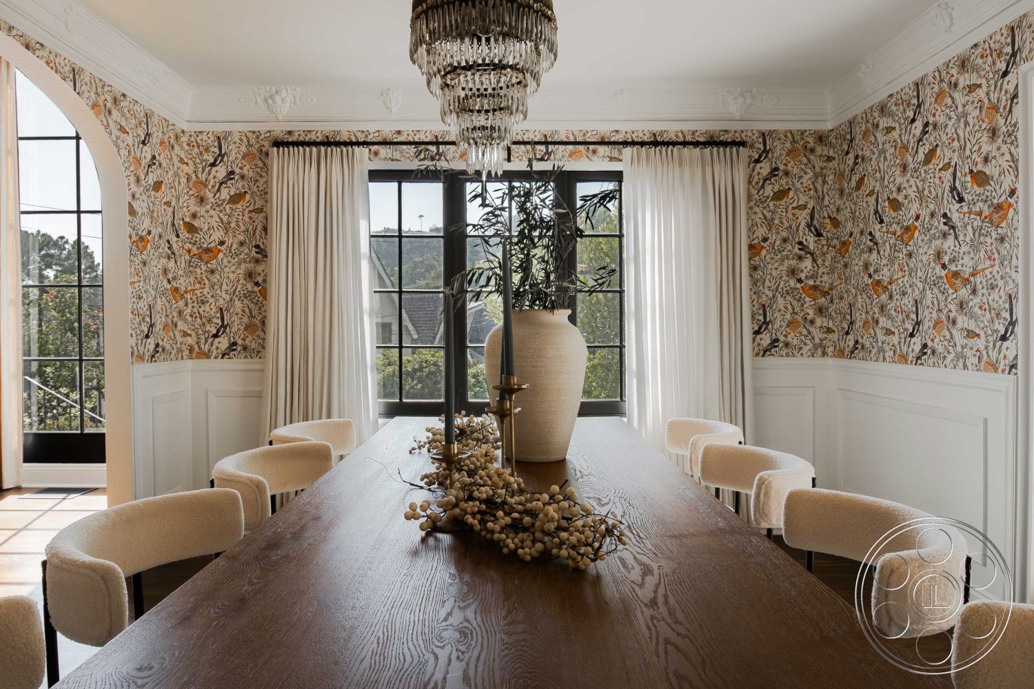 Spanish 166 - upholstered dining chairs, dark wood dining table, large black framed windows, natural light-filled space, interior dining room, beige fabric dining chair, floral wallpaper with orange birds, white wainscoting wall treatment, crystal chandelier lighting, floor-to-ceiling sheer curtains, ceramic vase on dining table, textured centerpiece with dried flowers, solid wood ceiling trim, open layout dining area