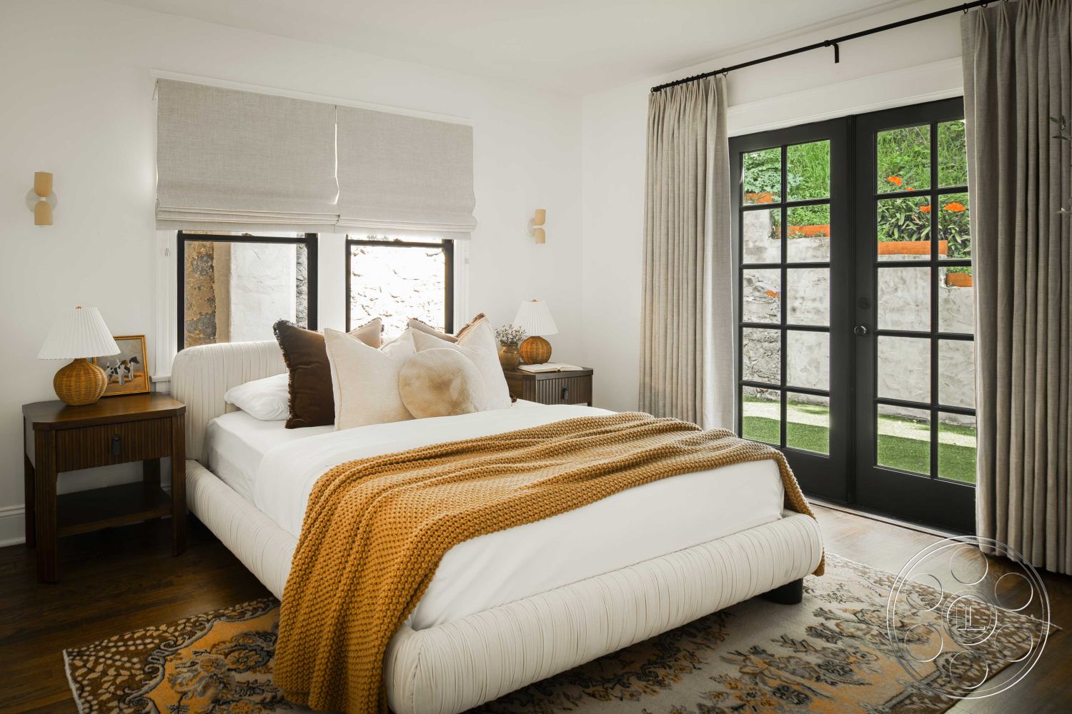 Spanish 166 - decorative throw pillows, decorative wall sconces, textured fabric curtains, interior bedroom space, hardwood bedroom floors, minimalist decor style, white painted walls, exterior garden view, linen roller shades, black framed French doors, cream-colored upholstered bed, wood nightstand with lamp, rug with intricate pattern, soft warm lighting, cozy bedding arrangement, plant accent near window, natural landscaping outside