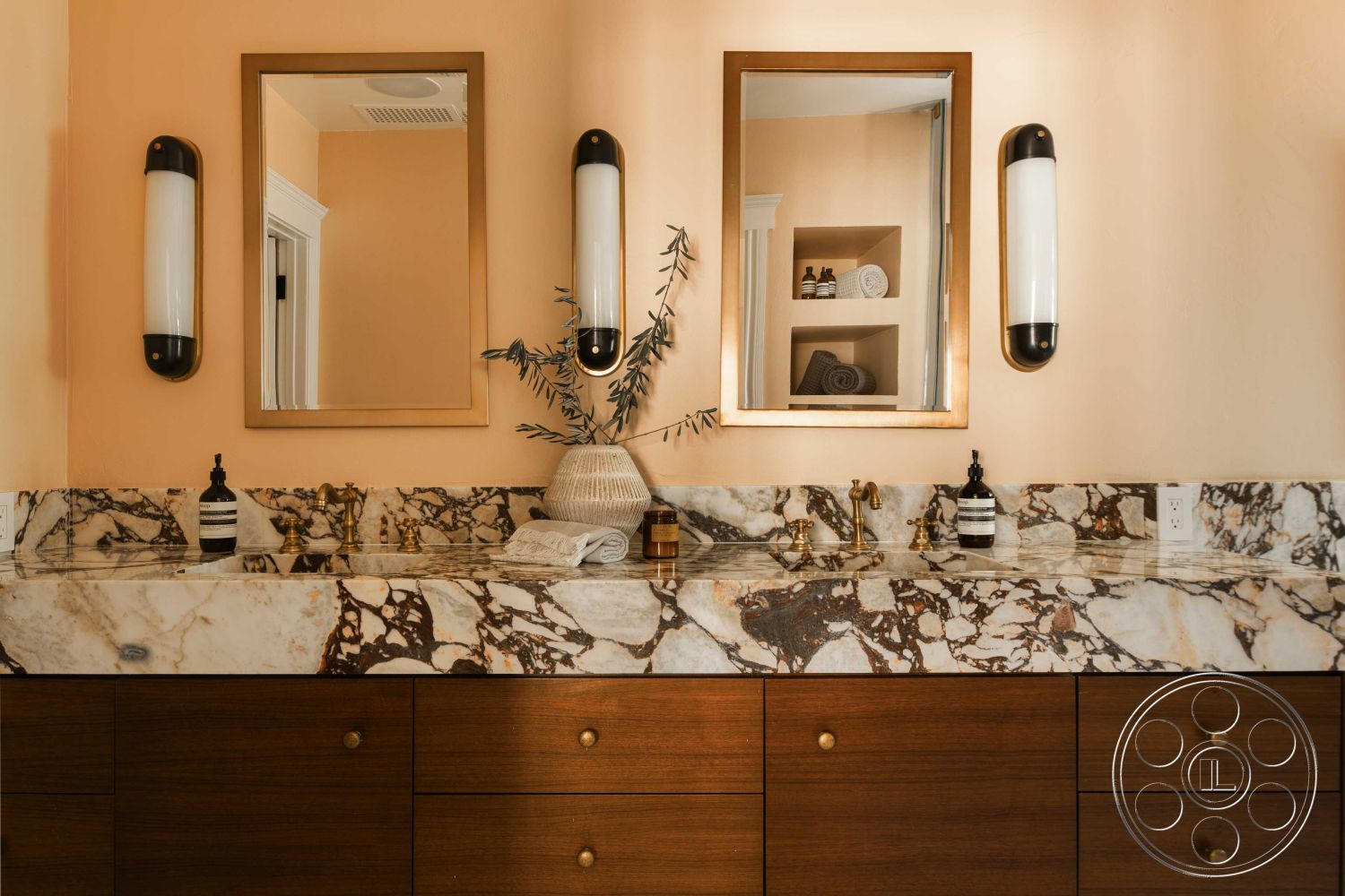 Spanish 166 - interior minimalist decor, ensuite, interior marble bathroom countertop, interior brown wood vanity cabinets, interior brass faucet fixtures, interior wall-mounted light sconces, interior framed bathroom mirrors, interior peach painted walls, interior decorative plant accent, interior recessed storage niche, interior soft towel display, interior modern lighting design, interior textured wash basin area