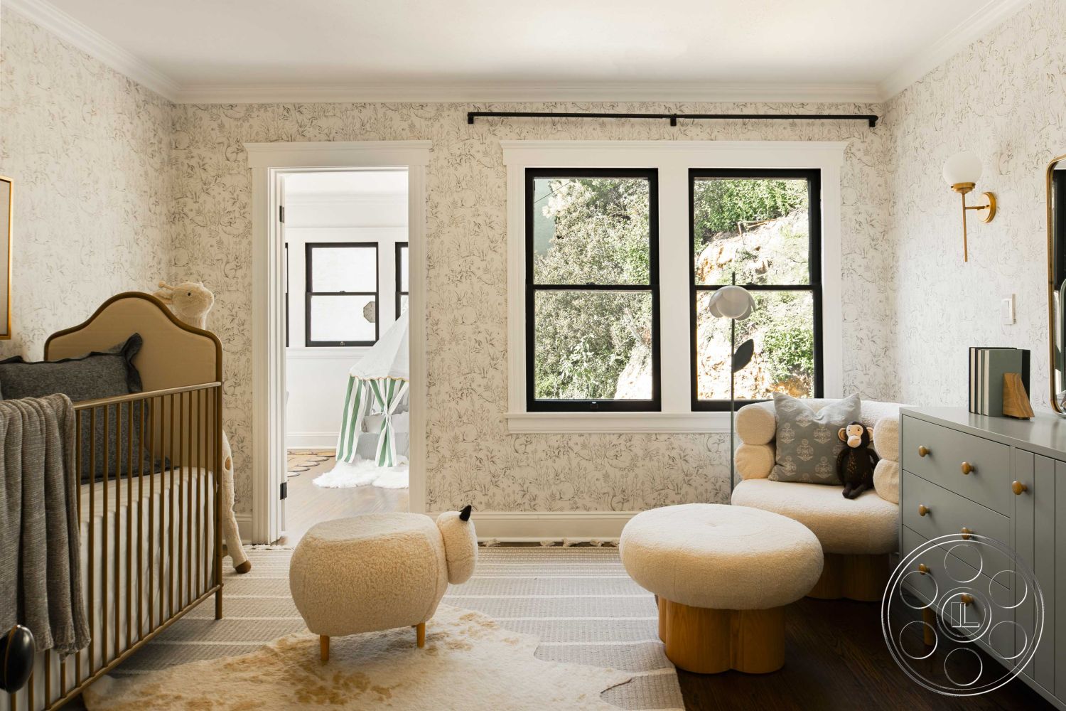 Spanish 166 - baby room, hardwood flooring, textured area rug, soft color palette, decorative accent pillows, white crown molding, interior nursery with floral wallpaper, exposed window frames, bronze wall sconce lighting, golden wood crib, neutral toned bedding, plush upholstered ottoman, modern grey dresser with wooden hardware, black frame windows with landscape view