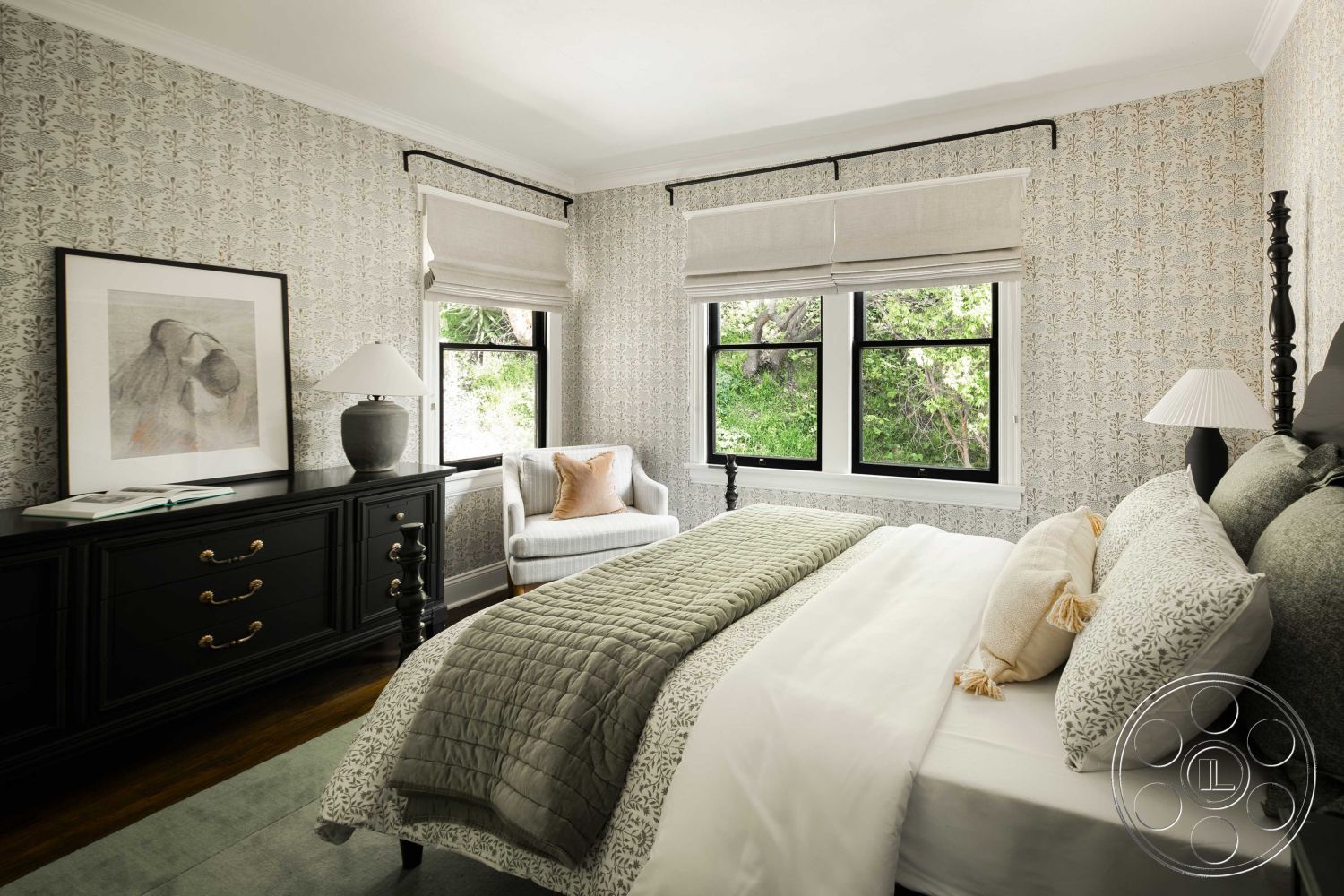 Spanish 166 - textured wallpaper, neutral color scheme, large framed artwork, natural light from windows, hardwood bedroom floors, plush gray bedding, black wooden dresser, black cabinet with gold hardware, beige decorative pillows, two-paned black windows, roman shades window treatment, green accent throw blanket, cozy reading nook with chair, four post bed