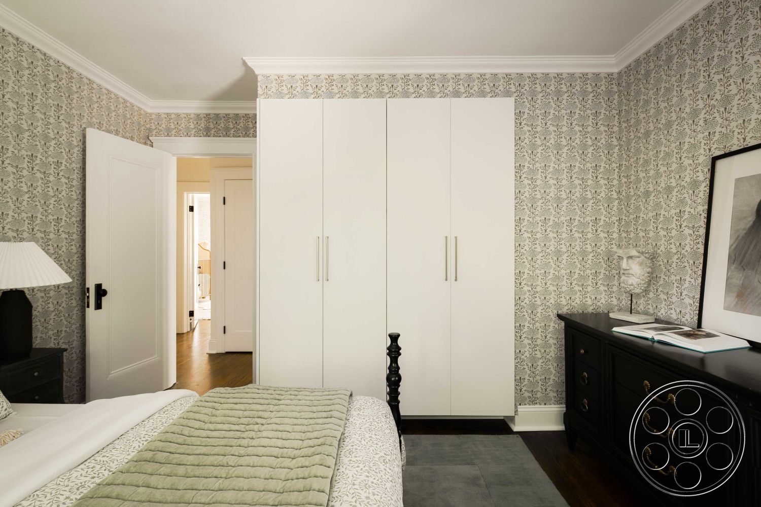 Spanish 166 - neutral color palette, minimalist interior design, hardwood bedroom floors, crown molding details, wallpapered accent wall, white modern closet doors, black dresser with brass hardware, textured bedding with green accents, classic table lamp on nightstand, contemporary style bedroom, styled art piece on dresser, elegant bedside decor, open doorway to hallway, natural light from nearby window