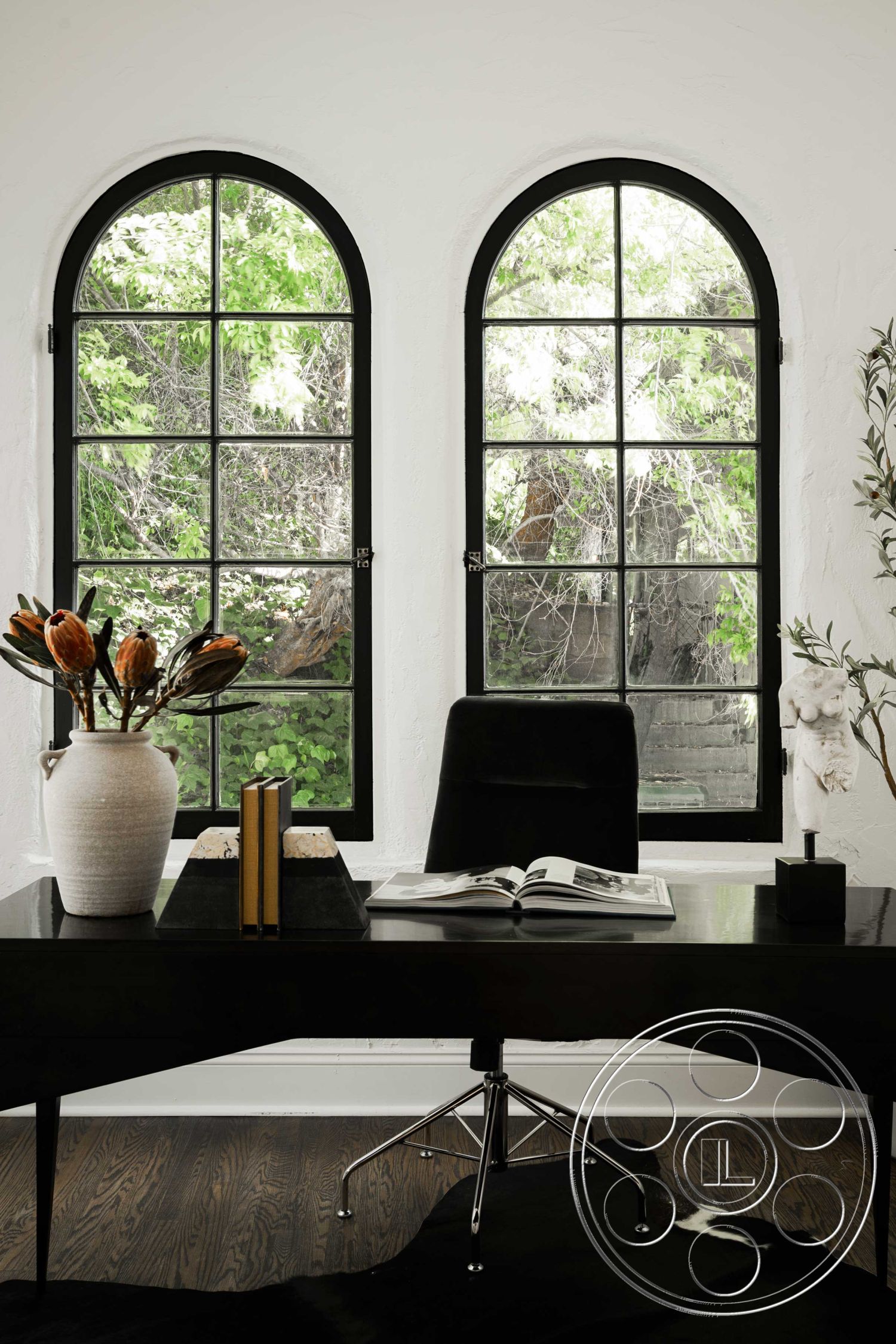 Spanish 166 - natural lighting, wooden flooring, neutral color scheme, interior office space, black glass top desk, armless black office chair, textured white plaster walls, arched black metal windows, green leafy outdoor view, modern decorative vase, minimalist decor aesthetics, books arranged on desk, cowhide area rug