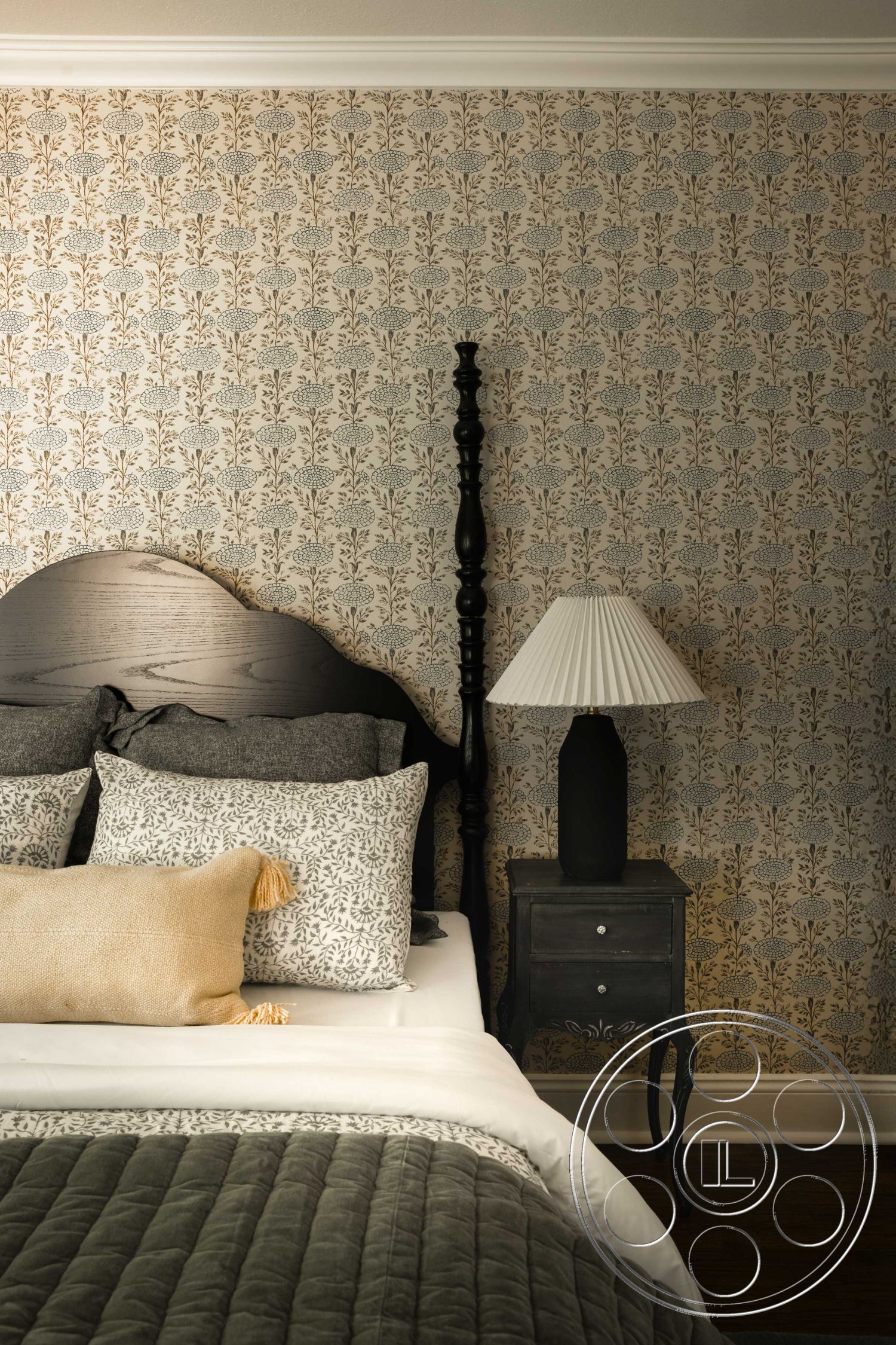 Spanish 166 - neutral color scheme, dark hardwood bedroom floors, cozy bedroom setting, floral wallpaper accent wall, black wooden bed frame, luxury bedding with decorative pillows, plaid patterned throw blanket, woven beige accent pillow, crown molding ceiling detail, black nightstand with drawer, table lamp with black base and white shade