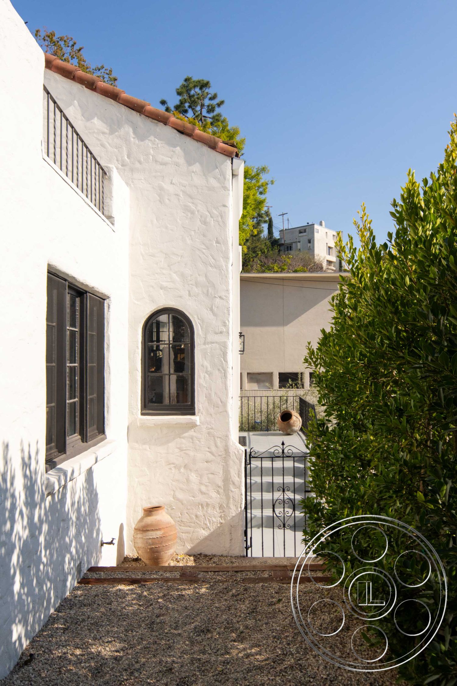 Spanish 166 - city skyline view, arched window, terra cotta roof tiles, gravel path, ceramic planter, wooden gate, exterior white stucco walls, urban backyard setting, green hedge landscaping, southwestern architectural style
