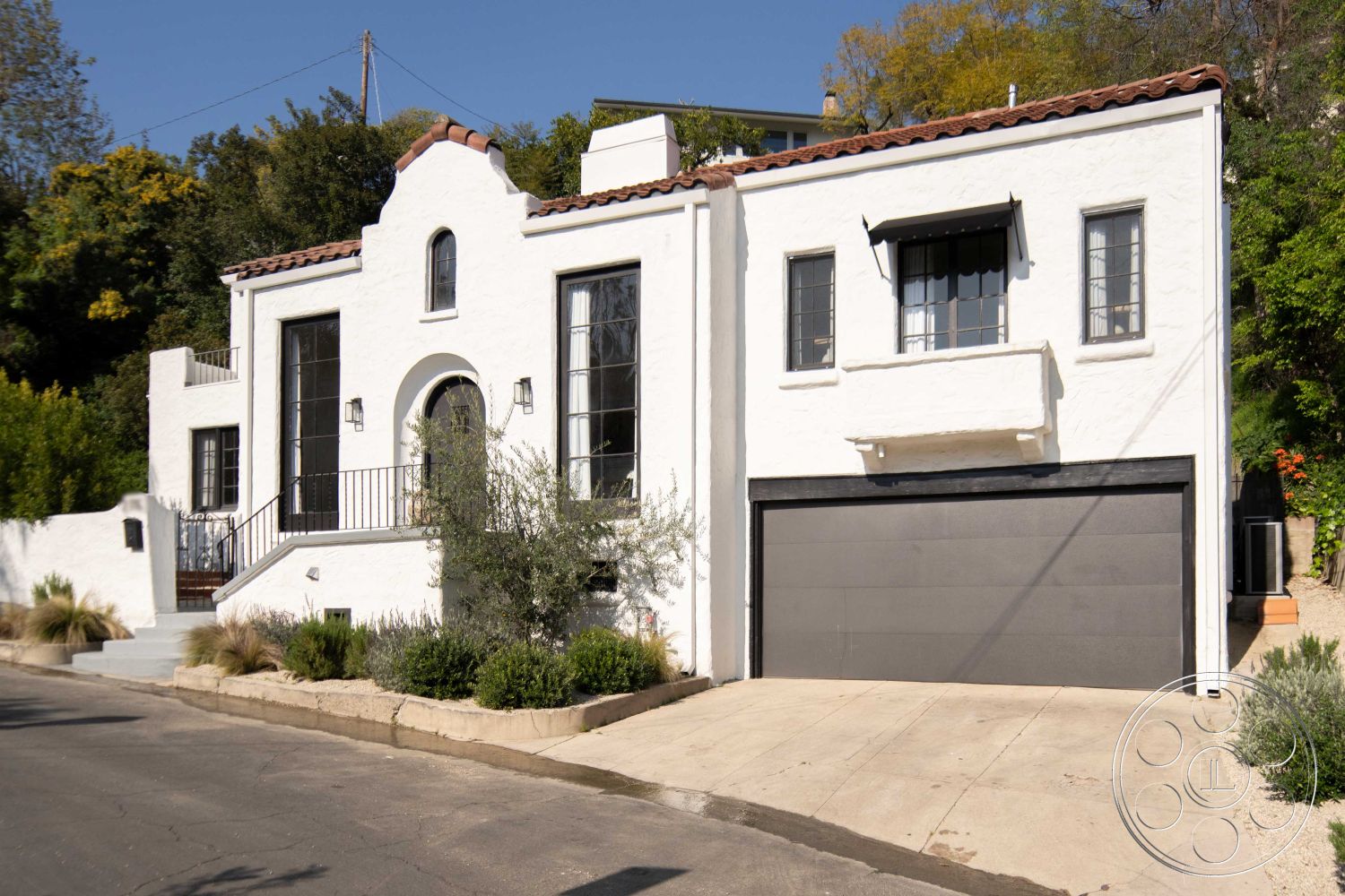 Spanish 166 - 2 car garage, concrete driveway, modern garage door, black window frames, landscape greenery, arched entryway, exterior stucco walls, Spanish tile roof, terraced landscaping, ornamental railing, slope-side location, driveway with gravel detailing, elevated porch steps, rounded exterior edges, large front windows, balcony overhang, exterior decorative wall sconces