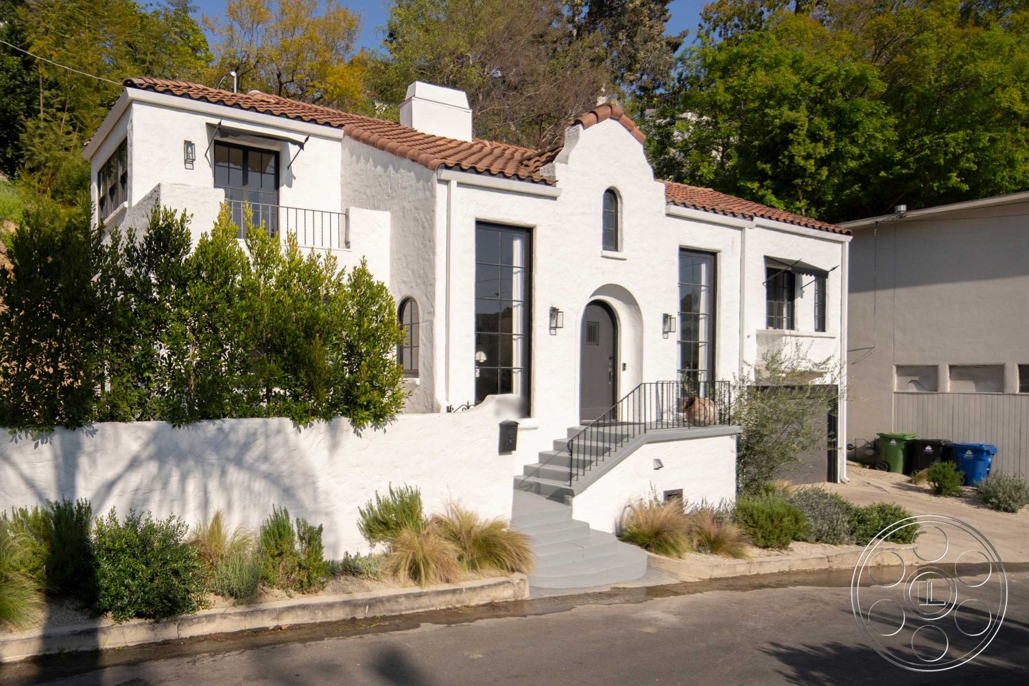 Spanish 166 - red tile roof, contemporary outdoor lighting, urban hillside location, exterior stucco walls, landscaped front garden, Spanish architecture, modern Mediterranean architecture, large black framed windows, low-maintenance shrubs, white plastered walls, iron railing staircase, arched front entryway, curved pathway leading to entrance, minimalist driveway design, elevated home facade, textured plaster surface, symmetrical building design, curbside
