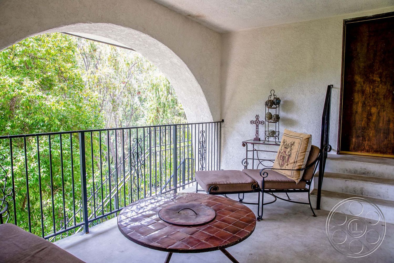 Medit 31 - patio seating area, rustic aesthetic, wrought iron railings, stucco wall finish, lush green landscaping view, exterior balcony space, arched balcony entryway, terracotta tile table, concrete floor patio, wooden bench with cushions, outdoor seating with table, decorative metal basket stand, soft beige upholstery, serene nature setting
