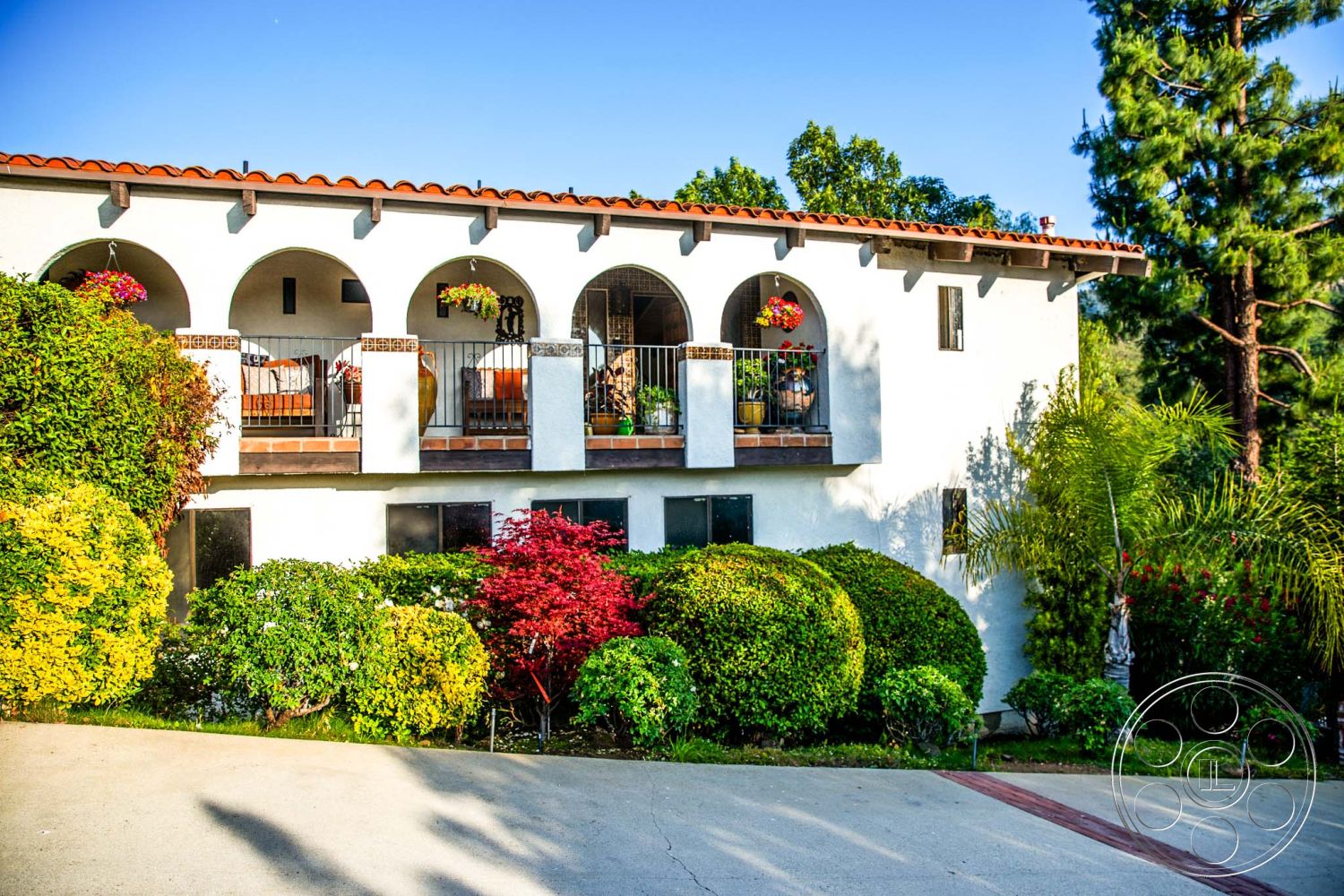 Medit 31 - mountain backdrop, Mediterranean architectural style, exterior white stucco walls, urban hillside setting, red terracotta roof tiles, arched wooden balcony railings, hanging flower pots, landscaped garden with ornamental shrubs, circular driveway with concrete surface, brick driveway border, rectangular windows with black frames, arched doorways leading to balcony, lush green surrounding vegetation