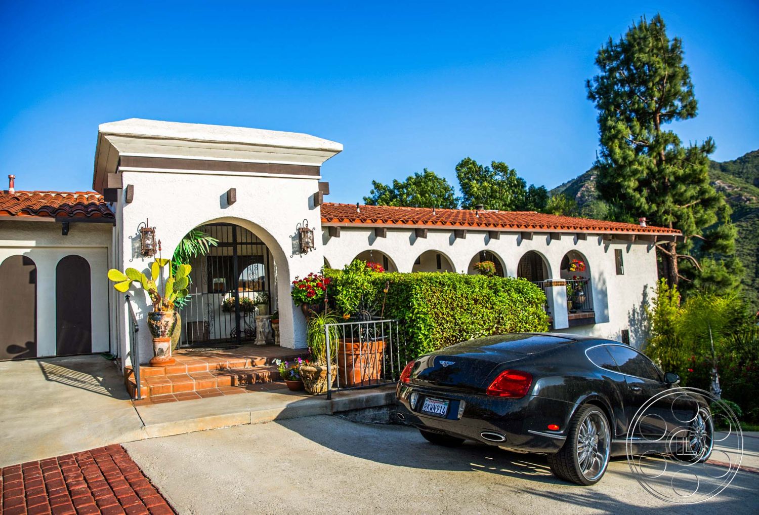 Medit 31 - paver driveway, slate stone pathway, curved driveway, mountain backdrop, red tile roof, arched doorways, Mediterranean architectural style, stucco exterior walls, lush greenery setting, terracotta patio tiles, exterior wrought iron railings, decorative outdoor lanterns, landscaped garden planters, potted cacti accents, open courtyard entry