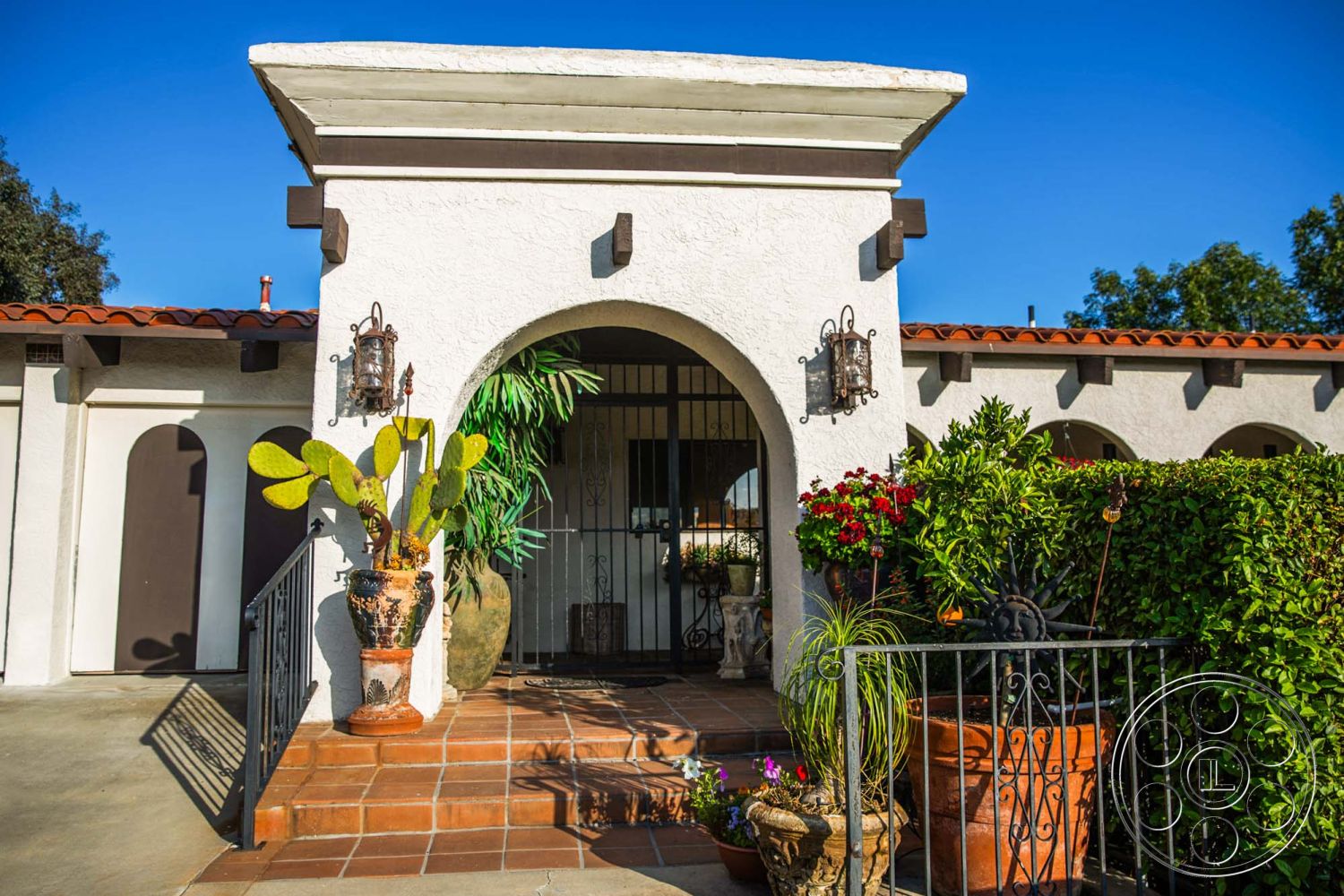Medit 31 - desert landscaping, red tile roof, textured plaster walls, arched entryway, Mediterranean architectural style, decorative wall lanterns, wrought iron railings, stucco exterior walls, landscaped front garden, exterior patio area, exposed wooden ceiling beams, natural light entry, terracotta plant pots, brick paver entry steps, ornamental garden planters, outdoor potted plants, curved arch entryway, accessible front doorway, interior courtyard access