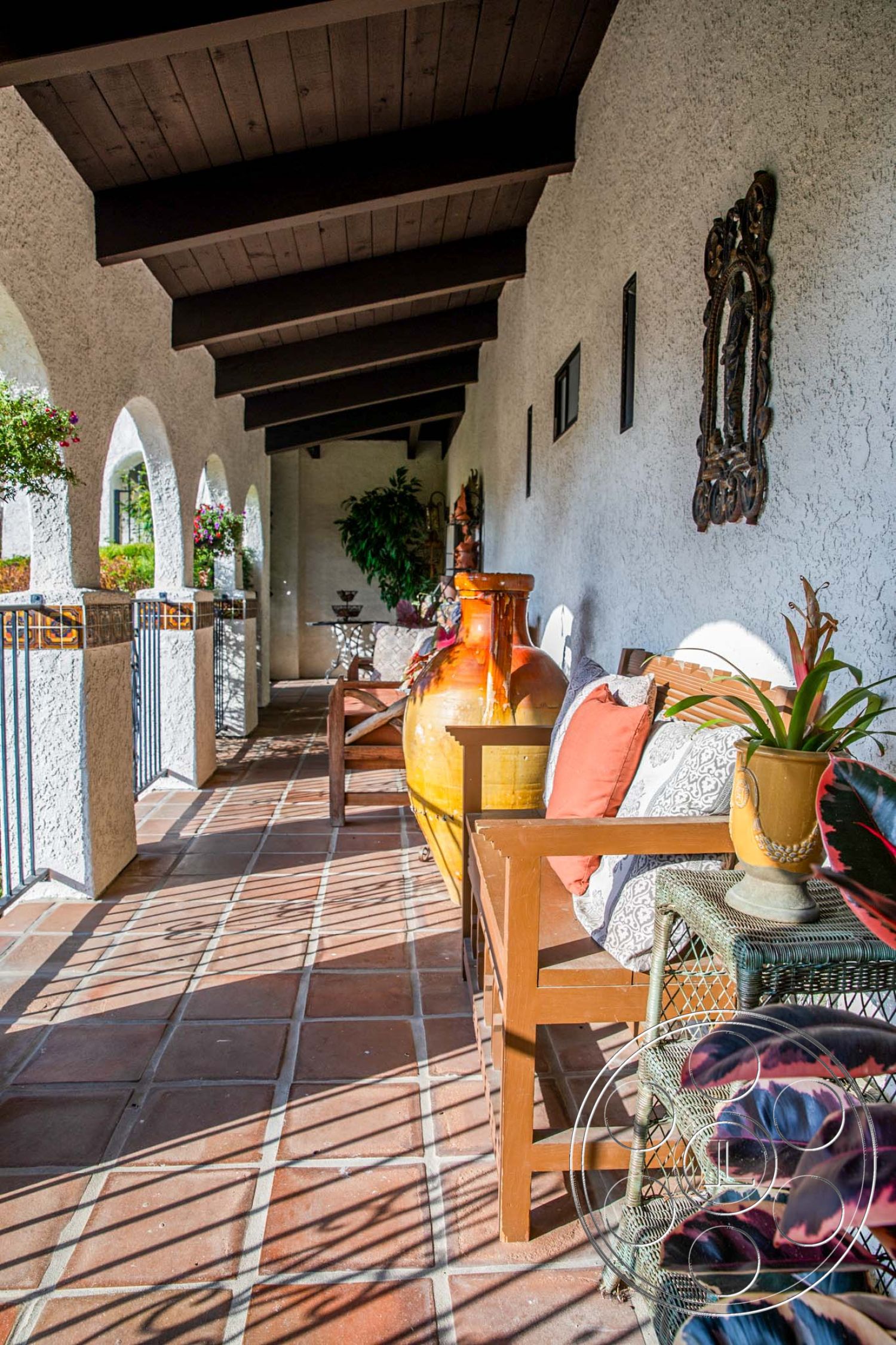 Medit 31 - artistic wall decor, arched doorways, Spanish-style architecture, decorative ceramic vase, textured wall detail, exposed wood beam ceiling, exterior plaster walls, red clay tile flooring, rustic patio area, woven outdoor chair, vibrant outdoor cushions, greenery accents with potted plants, shadow play from railing, open-air porch setting, bright sunny ambiance