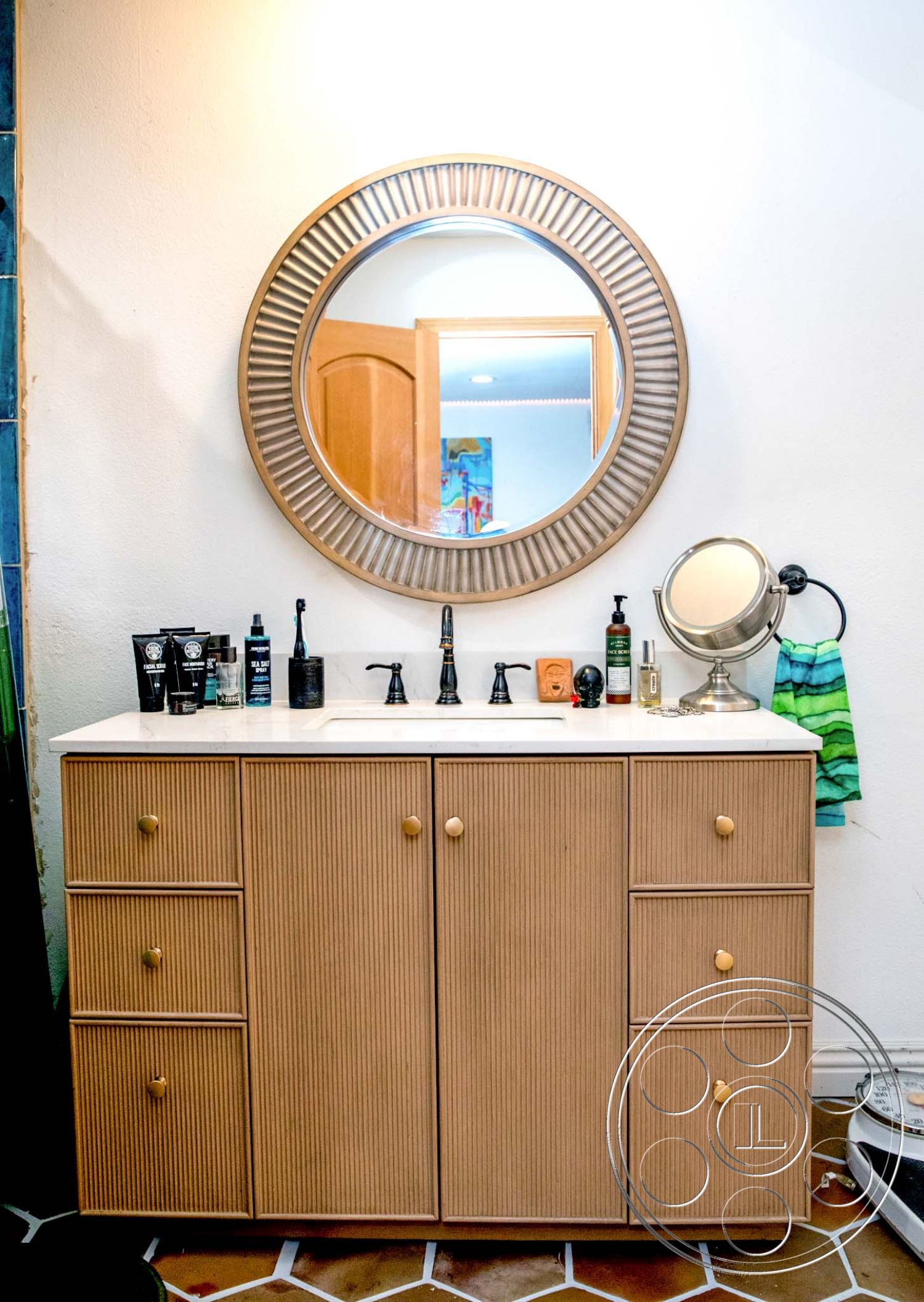 Medit 31 - textured wall paneling, hexagonal tile flooring, white solid surface countertop, gold cabinet hardware, brass faucet fixtures, interior bathroom vanity, laminate wood grain cabinet doors, round decorative wall mirror, metal makeup mirror, colorful wall art in background, towel hanging on hook, bathroom countertop accessories