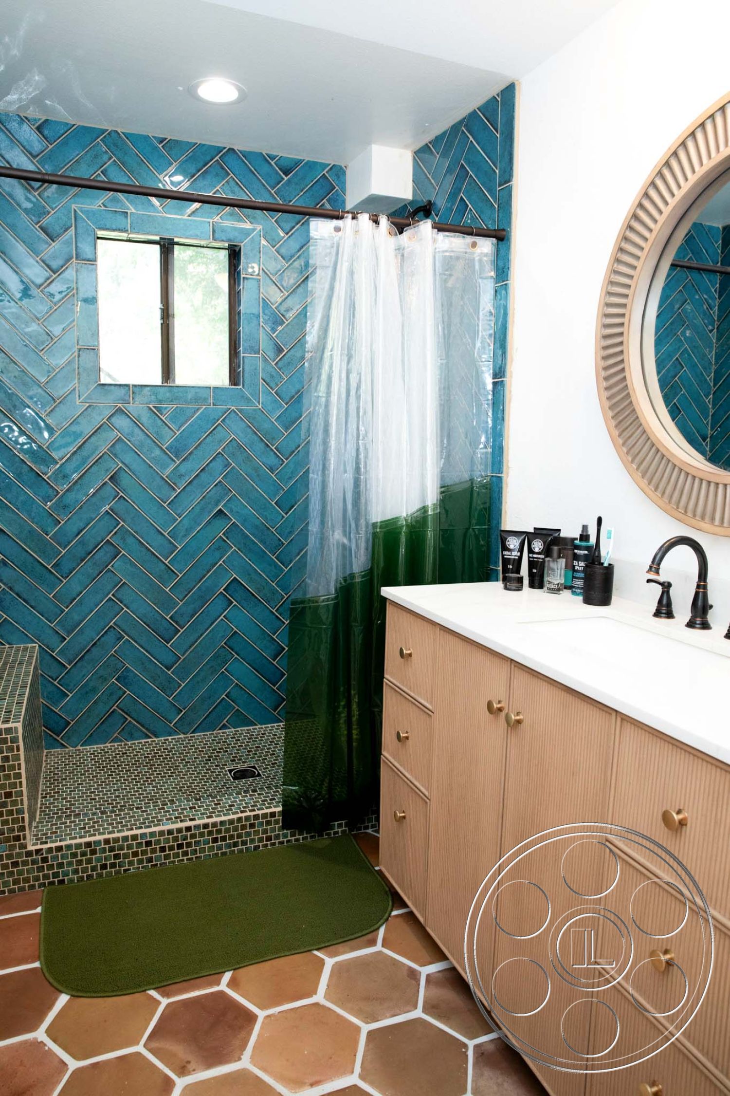 Medit 31 - textured plaster ceiling, contemporary aesthetics, natural light from window, minimalist design with clean lines, interior bathroom with teal herringbone tile walls, hexagonal terracotta floor tiles, glass shower curtain with gradient color, modern wooden bathroom vanity with gold hardware, woven green bath mat, framed round mirror, mosaic tiled shower floor, exposed plumbing fixtures in matte black