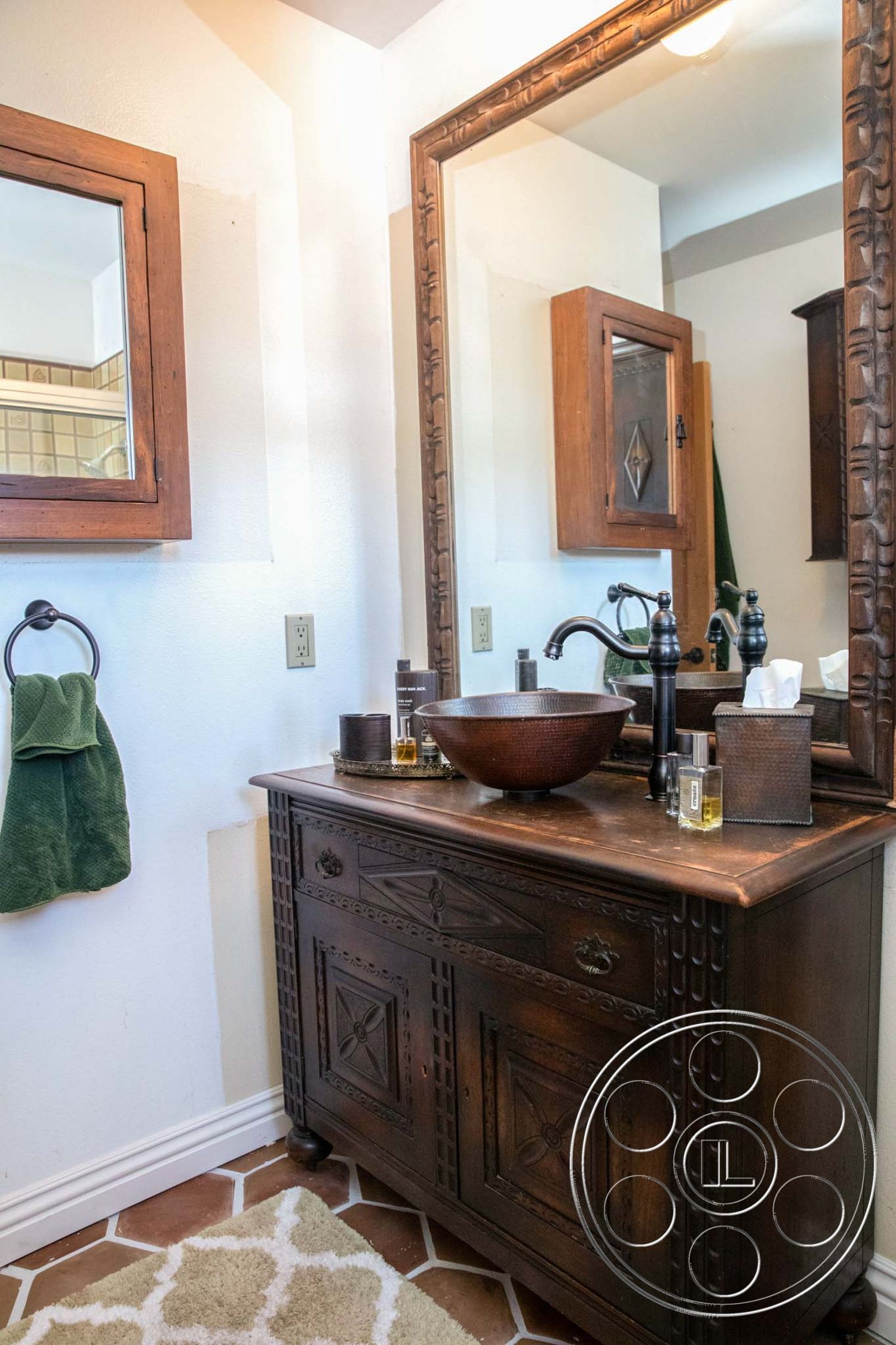 Medit 31 - interior bathroom, white painted walls, wood bathroom vanity, brass faucet fixtures, carved wood mirror frame, oval bronze sink, green towel hanging, textured ceramic floor tiles, earth-toned bathroom flooring, solid wood cabinetry, decorative cabinet accents, vintage-style bathroom, rustic bathroom design