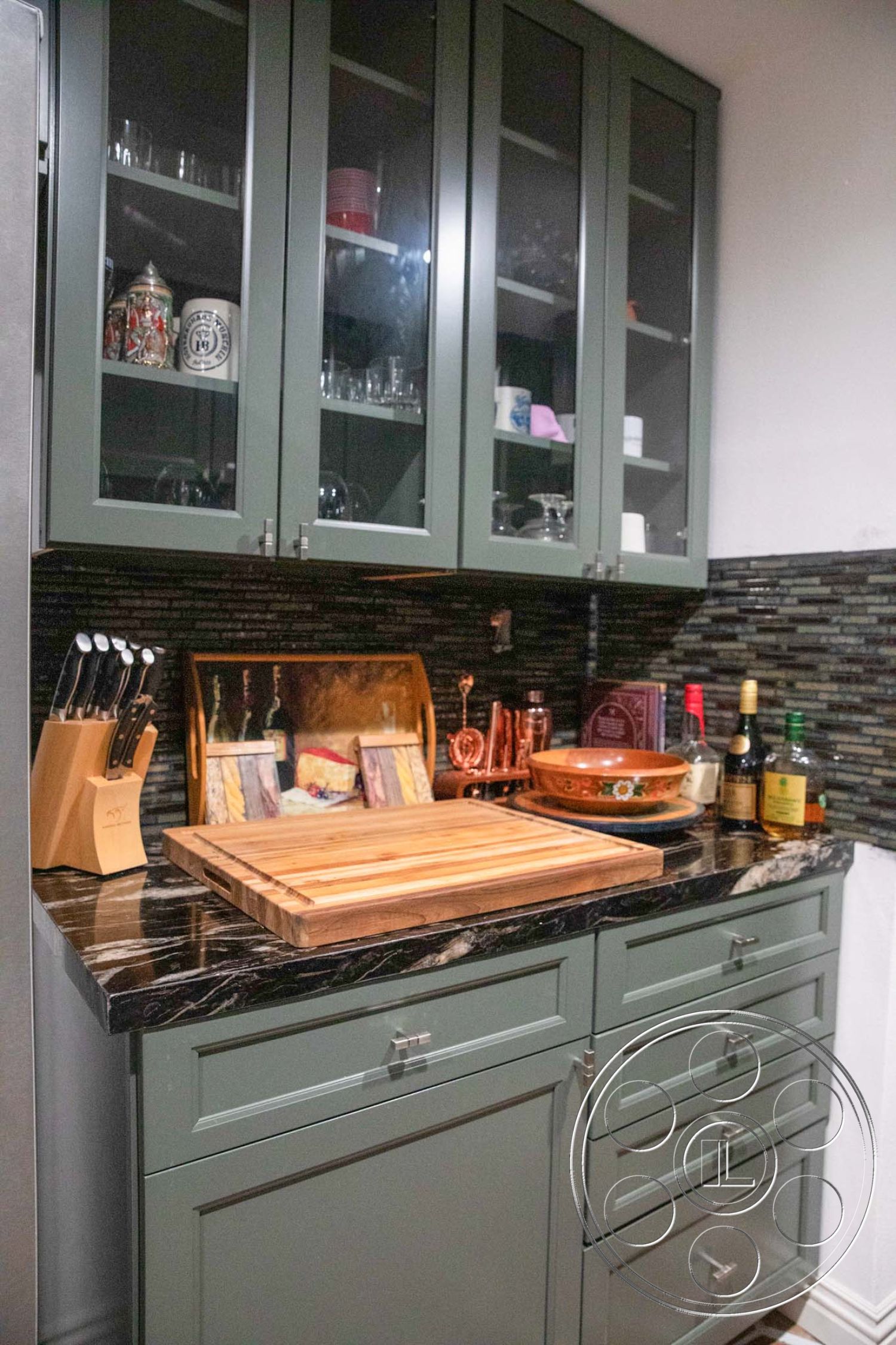 Medit 31 - wood cutting board, glassware storage, stainless steel refrigerator, textured tile backsplash, functional kitchen workspace, interior kitchen with green cabinetry, marble countertop with dark veining, glass-front kitchen cabinets, modern kitchen accessories, open shelving display
