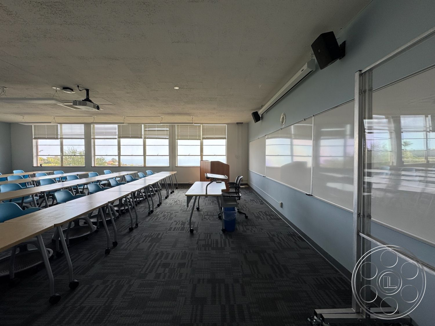 School 1 - modern lighting fixtures, acoustic ceiling tiles, textured carpet flooring, large windows with blinds, interior classroom, solid wood teacher desk, steel-framed chairs, whiteboard wall panels, rectangular classroom layout, neutral pastel wall color, overhead projector, open spacious environment, expansive natural light, wireless speaker system, collaborative seating arrangement, functional learning space