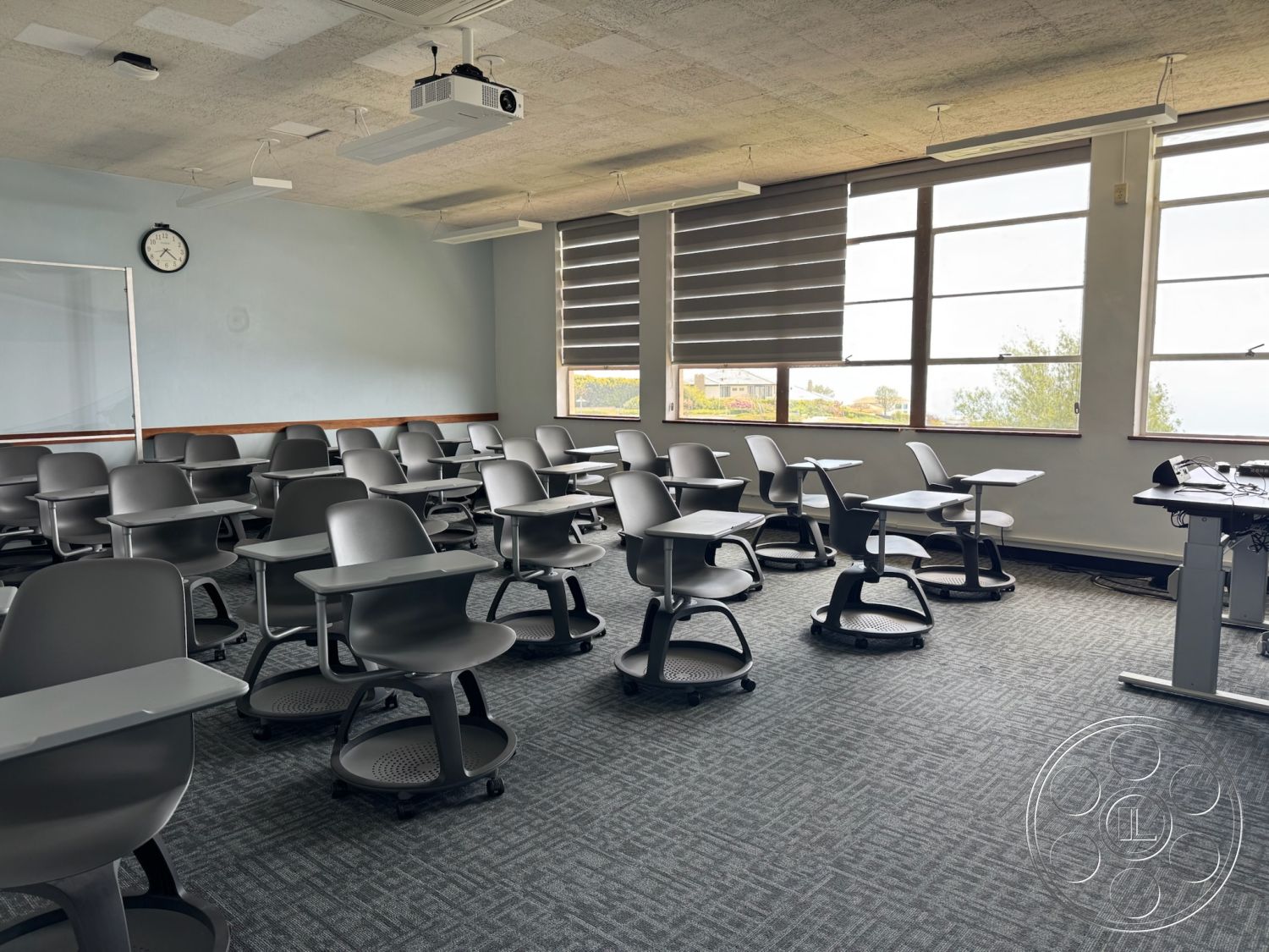 School 1 - natural light from windows, wall clock, acoustic ceiling tiles, textured carpet flooring, interior classroom space, gray upholstered classroom chairs, roll-around chairs with attached desks, large windows with horizontal blinds, wood accent trim along walls, integrated projector system, open layout with flexible seating, modern educational environment, collaborative learning space