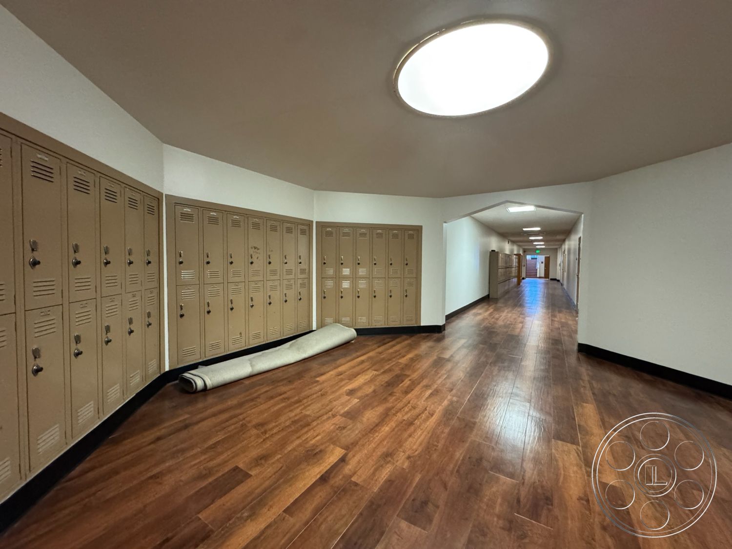 School 1 - neutral color palette, textured wall finish, curved wall design, circular ceiling light, white painted walls, industrial-style interior, vinyl wood plank flooring, beige metal lockers, rectangular hallway layout, rolled carpet runner, modern school corridor, multi-directional lighting