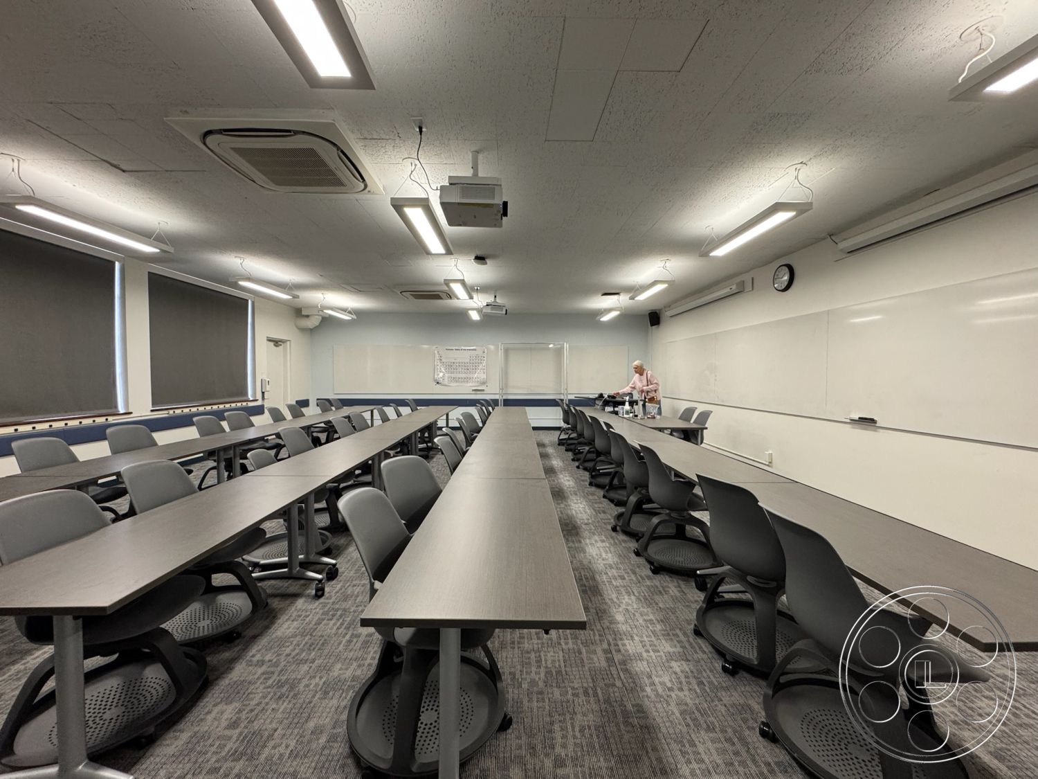 School 1 - black ergonomic chairs, wall-mounted projector, acoustic ceiling tiles, textured carpet flooring, large windows with blackout shades, interior classroom setting, fluorescent ceiling lighting, gray laminate tables, motion-sensor ceiling fan, white dry erase board, circular wall clock, white wall paint, exposed HVAC ductwork