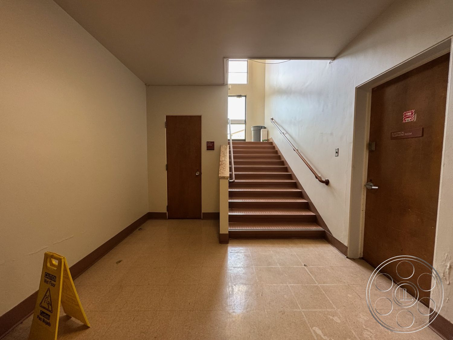 School 1 - brown wooden doors, minimalist architectural style, high ceiling with natural light, interior stairs with tiled flooring, vinyl stair treads, plain white painted walls, aluminum handrail, corner trash bin, caution wet floor sign, no decoration or art, functional public building design, simple pedestrian flow, utilitarian hallway
