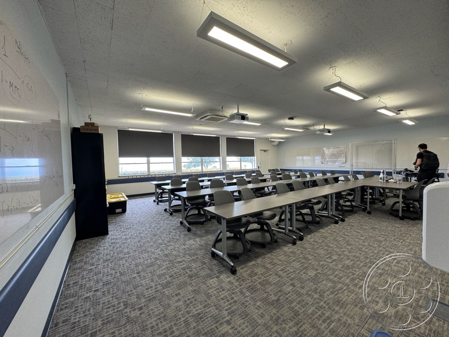 School 1 - open floor plan, acoustic ceiling tiles, interior classroom setting, carpeted flooring with geometric pattern, modern adjustable task chairs, long rectangular conference tables, large windows with blackout roller shades, whiteboard wall with mathematical equations, projector and screen setup, LED overhead lighting fixtures, neutral color palette with blue accent stripe, collaborative work space layout, air conditioning units mounted on ceiling, teacher's desk area, back wall storage unit for supplies