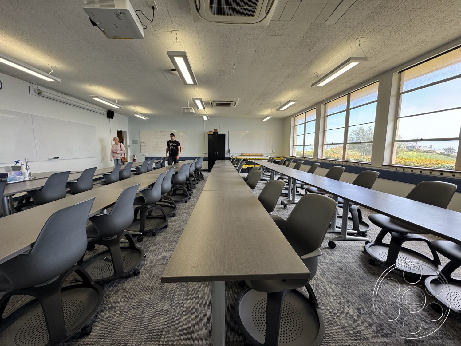 School 1 - collaborative workspace, ergonomic office chairs, acoustic ceiling tiles, accessible design features, large windows providing natural light, LED overhead lighting, interior classroom setting, modern classroom design, gray laminate tabletop desks, multiple rows of desks, carpeted flooring with geometric patterns, whiteboard walls, projector mount, open learning environment, dual-purpose classroom layout