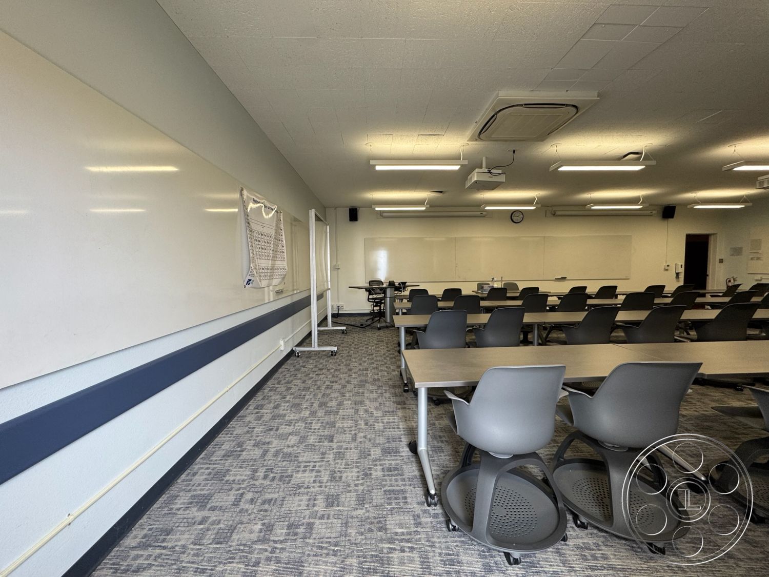 School 1 - grey carpet flooring, open floor layout, whiteboard wall panels, interior classroom space, textured acoustic tile ceiling, light wood conference tables, stackable plastic swivel chairs, blue accent stripe on wall, modern linear lighting fixtures, projector screen mount, wireless microphone setup, wall-mounted clock, collaborative learning environment