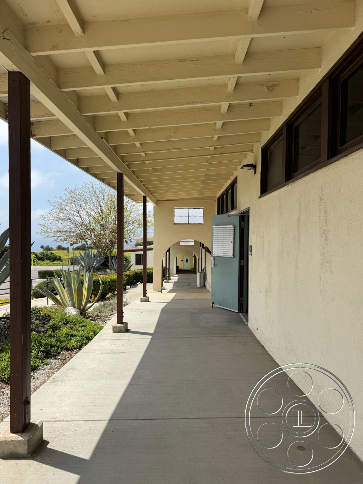 School 1 - outdoor landscaping, arched doorways, spacious corridor, sunny outdoor setting, concrete sidewalk, stucco wall finish, wood beam ceiling, exterior walkway, low-maintenance shrubbery, polished concrete surface, desert plantings, lightly textured wall surface, functional exterior lighting, horizontal window design, open-air passageway