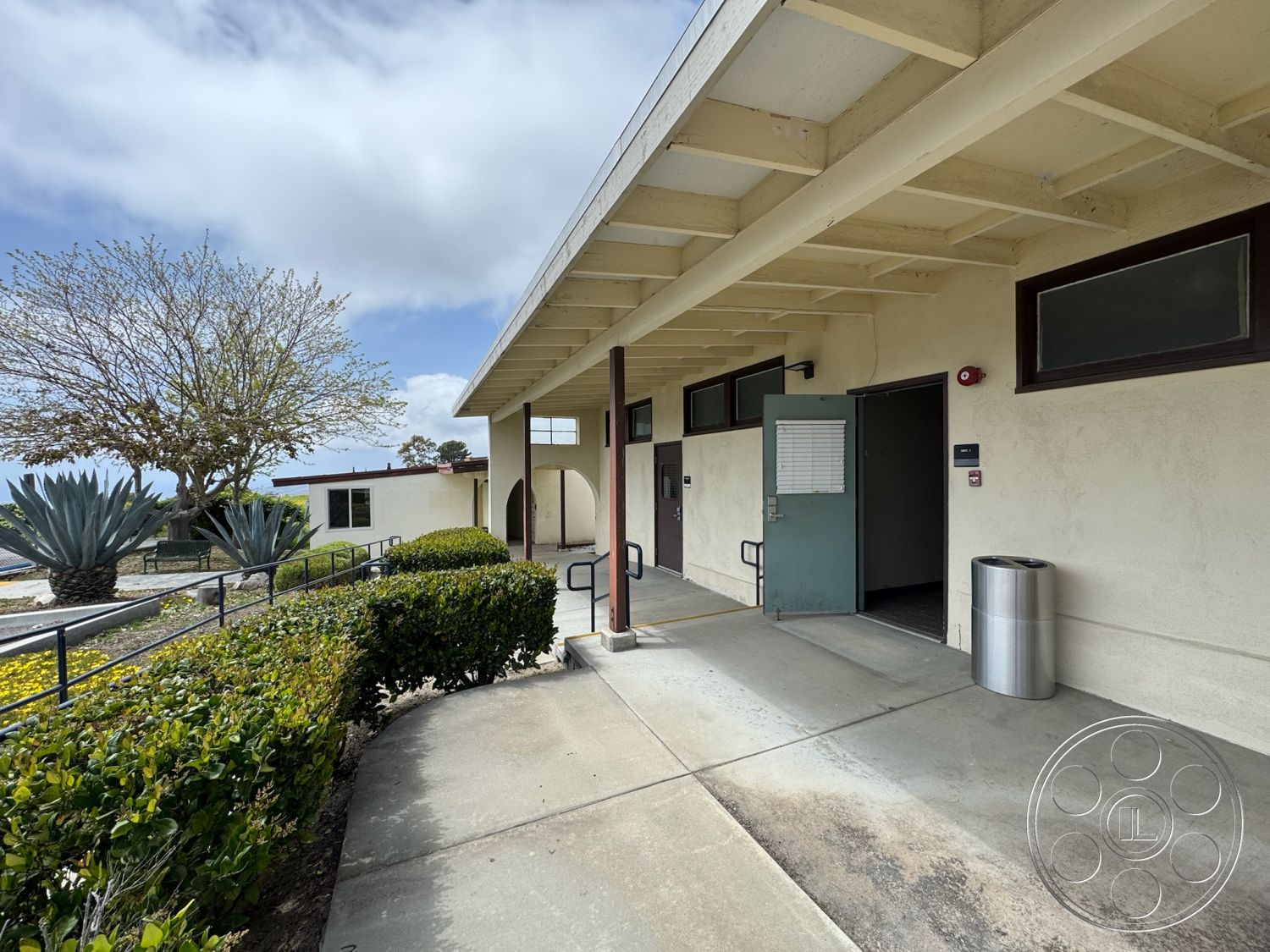 School 1 - exterior low-maintenance landscaping, exterior concrete walkway, simple geometric forms, exterior textured stucco walls, exterior concrete ramp, wooden support beams, light-colored concrete surfaces, flat roof architectural design, open entrance with glass door, modern entryway with accessibility features, surrounding drought-resistant plants, urban setting with distant trees, entry area with shaded overhang, well-defined architectural lines, integrated waste receptacle, exterior landscaped border, accessible exterior building entrance, natural light through clerestory windows