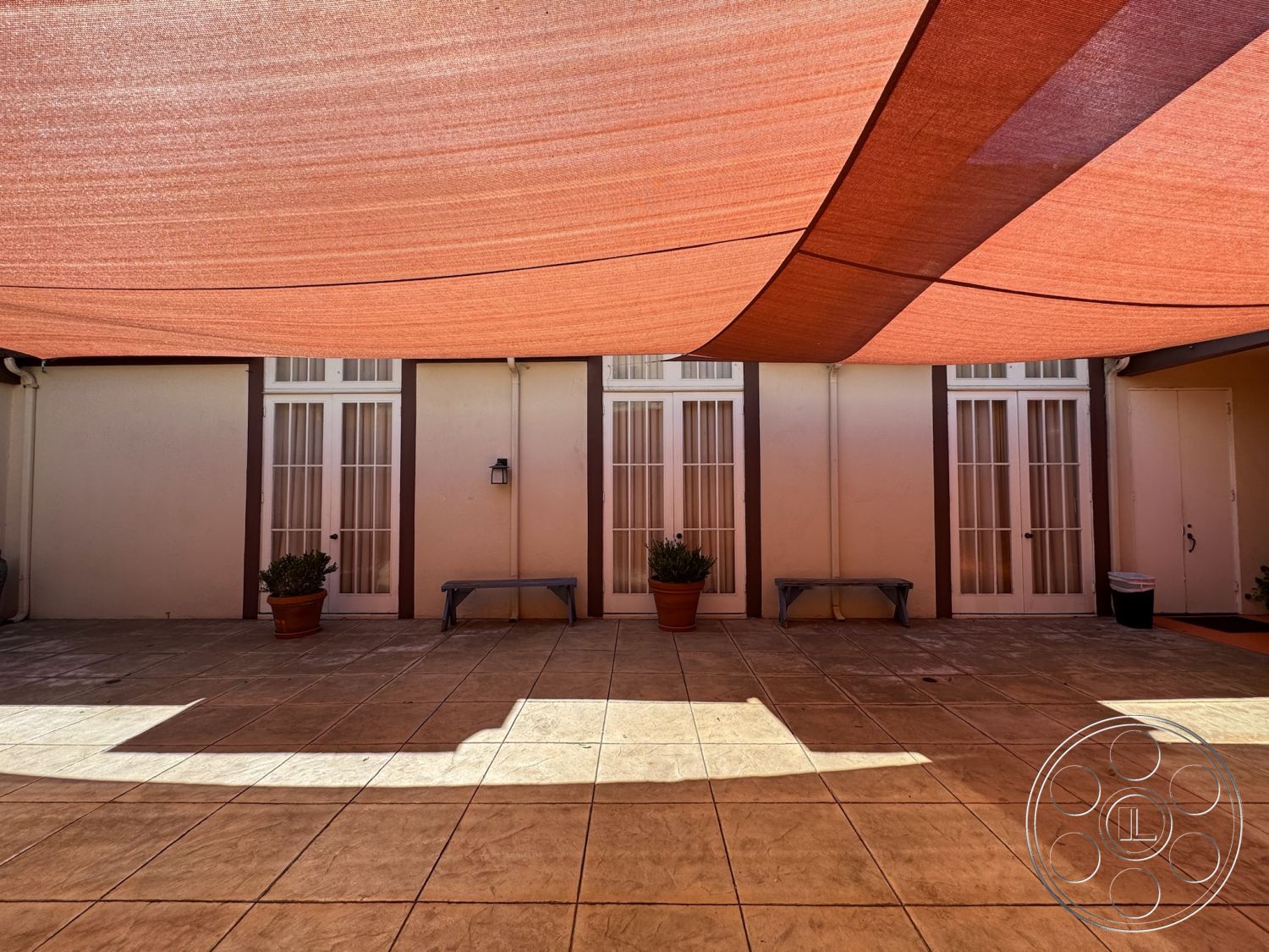 Spiritual Center - modern outdoor seating area, exterior patio flooring, stamped concrete patio floor, tan-colored concrete tiles, benches with wooden slats, overhead shade sail, modern glass door entrance, potted plants on patio, neutral wall treatment, city courtyard setting