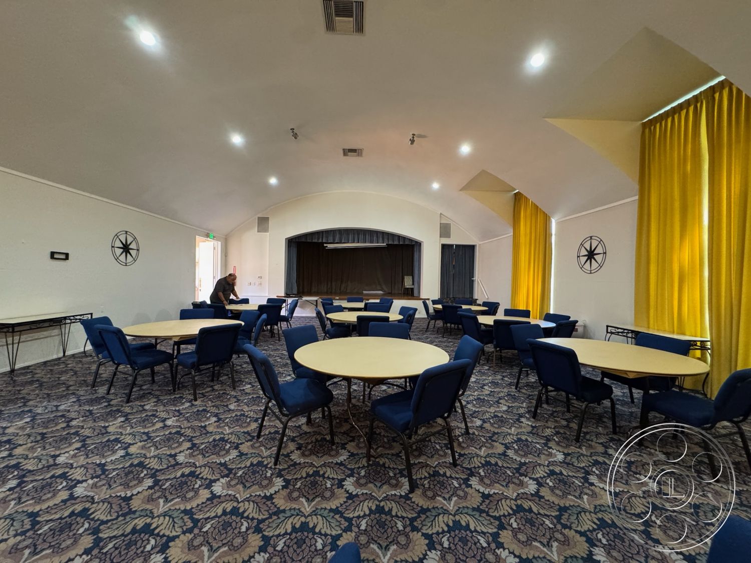 Spiritual Center - large windows, sconce lighting, interior space, carpet flooring, soft ambient lighting, multipurpose room, metal folding chairs, white painted walls, acoustic ceiling treatment, floral patterned carpet, round tables with blue upholstery, high ceiling with curved design, yellow fabric curtains, stage area with dark curtain, wall-mounted decor, coffee station area, entry door with clear glass panel