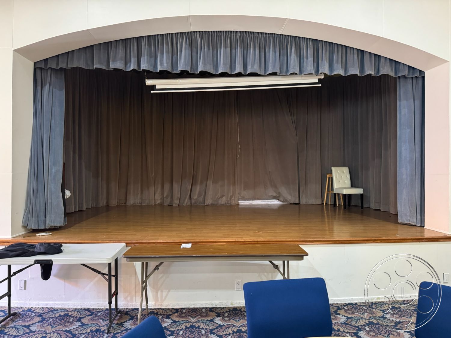 Spiritual Center - blue upholstered chairs, neutral painted walls, acoustic wall treatment, wood stage flooring, curved stage archway, blue curtain backdrop, folding table setup, lighting rig above stage, performing arts space interior, multi-purpose room design, theater stage area, wooden chair on stage, carpeted event floor, adjustable track lighting, seating arrangement for audience