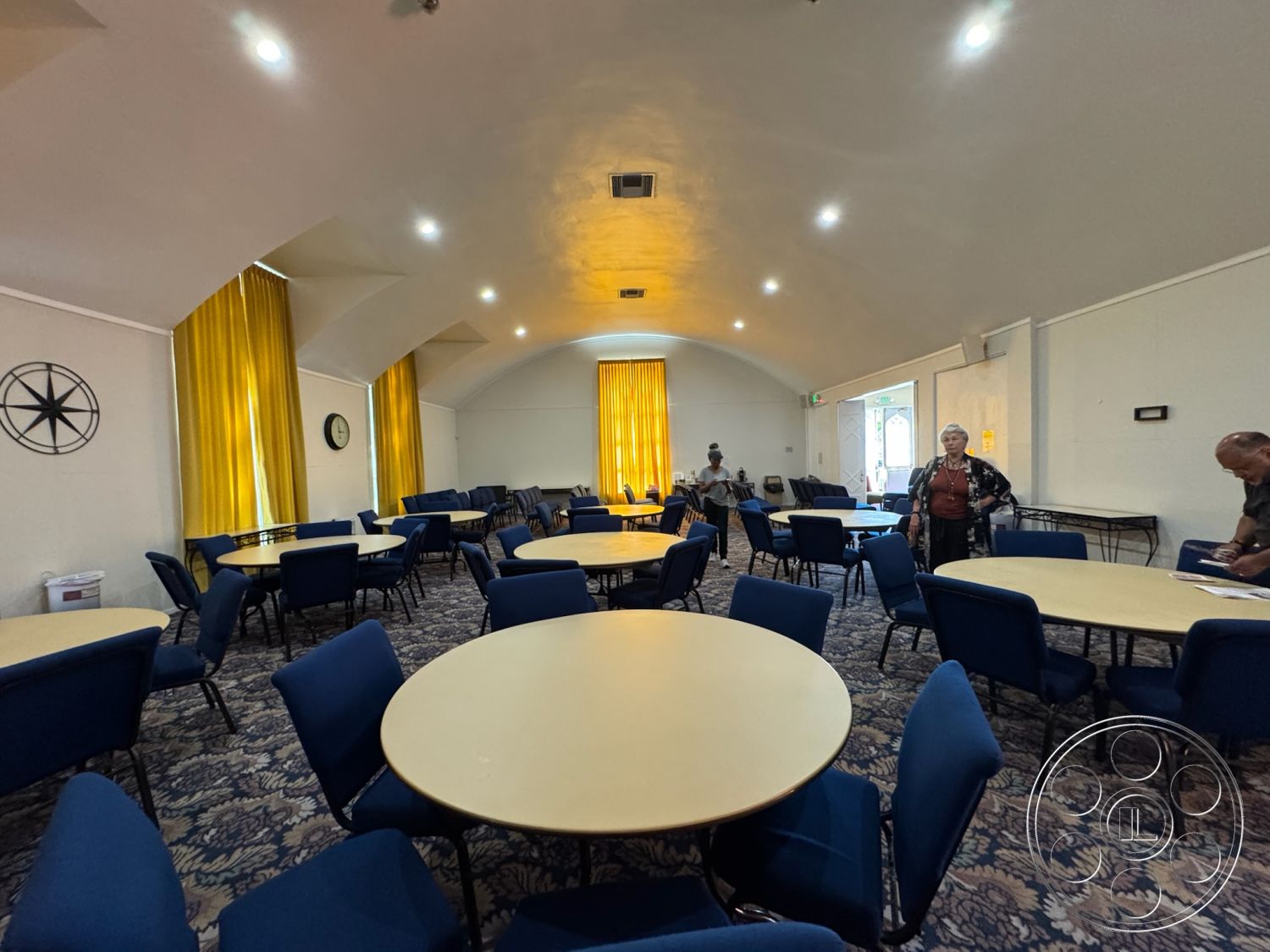 Spiritual Center - open floor plan, interior space, blue upholstered chairs, arched ceiling, decorative wall clock, soft carpeting, community gathering area, large entryway, acoustic ceiling tiles, neutral wall paint, ambient recessed lighting, round dining tables, window treatments with yellow curtains, accessible seating layout