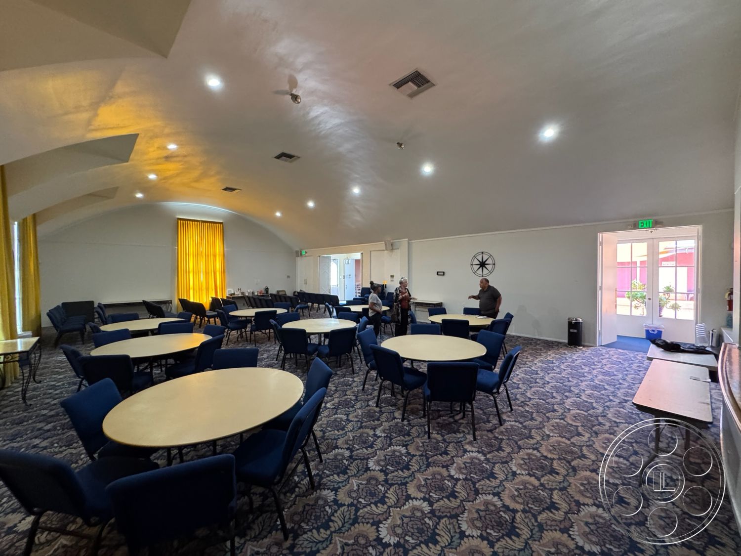 Spiritual Center - modern light fixtures, floor-to-ceiling windows, textured plaster walls, decorative wall clock, round wooden tables, acoustic ceiling panels, open layout space, double door entrance, interior multi-purpose room, arched ceiling design, dark blue upholstered chairs, yellow sheer curtains, patterned carpet flooring, wooden exit door