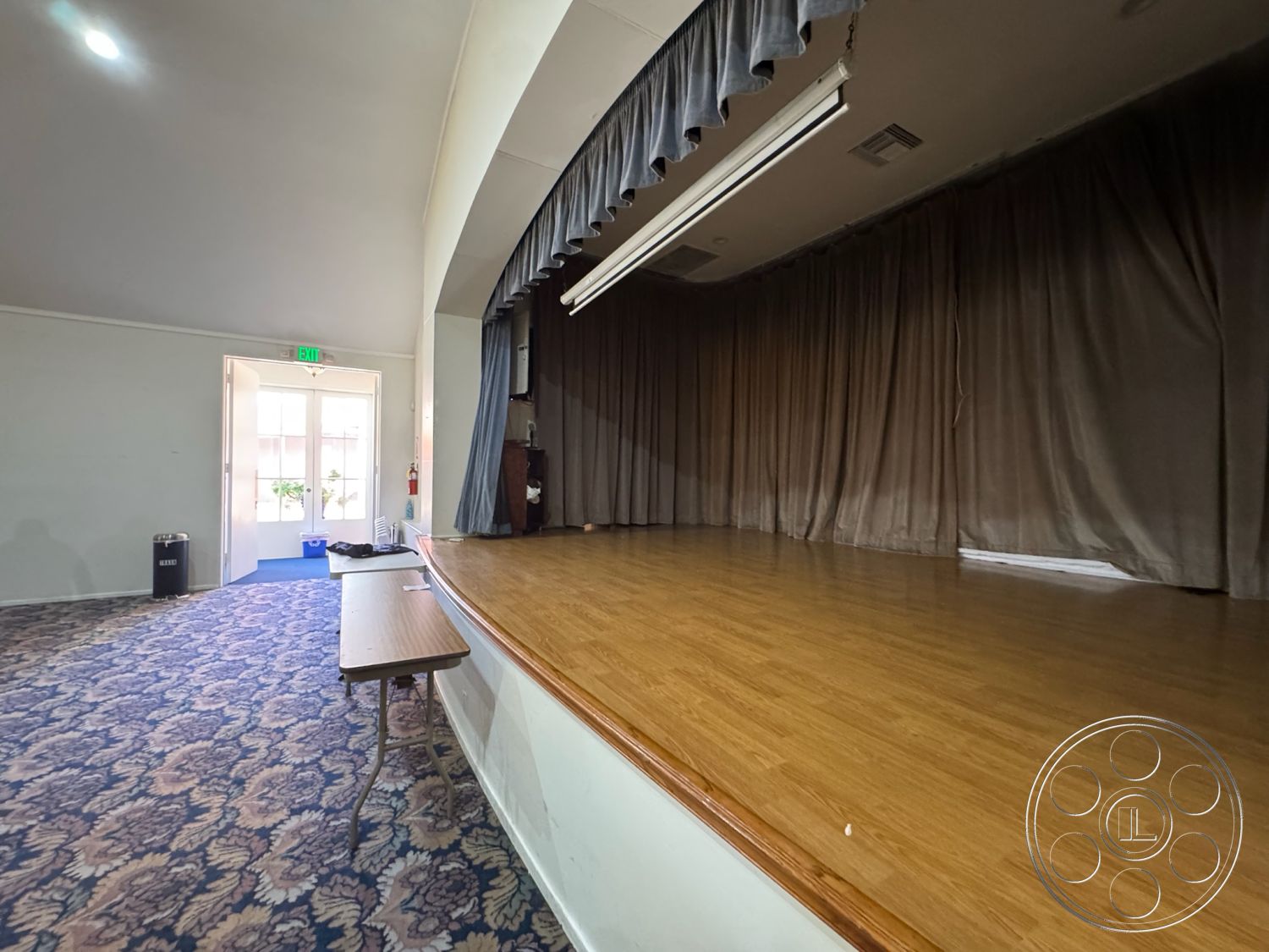 Spiritual Center - neutral color palette, exposed ceiling beams, double door entry, community gathering area, recessed ceiling lighting, open floor plan layout, flush-mount ceiling lights, hardwood stage flooring, curved stage edge, heavy fabric stage curtains, textured wallpaper walls, decorative carpet flooring, vintage-style stage backdrop, interior event space, multipurpose hall, acoustic curtain treatment, wooden table for events