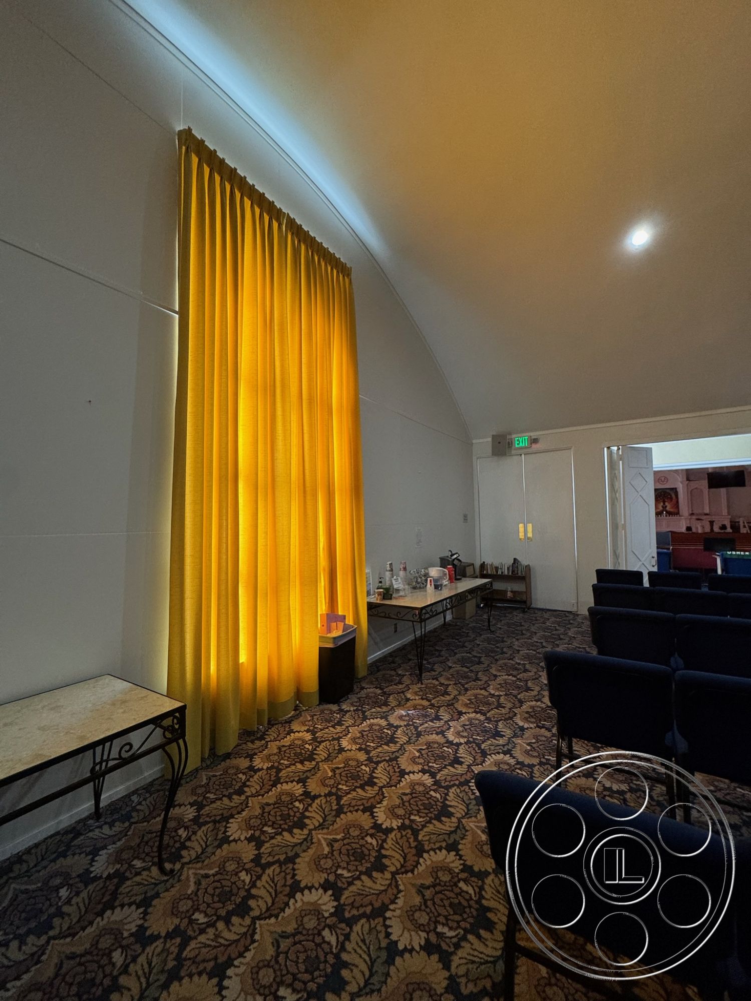 Spiritual Center - interior space, warm color palette, dim ambient lighting, recessed lighting fixtures, open room layout, arched ceiling design, yellow sheer curtains, decorative carpet flooring, intricate floral pattern carpet, simple black chairs, small serving table, stylish metal table legs, colorful decor accents, welcoming gathering space