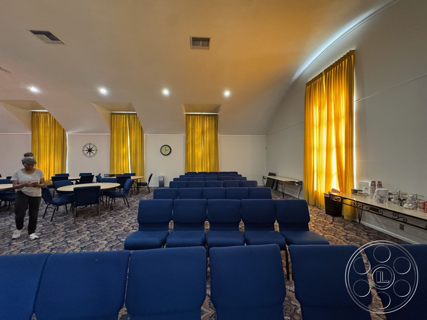 Spiritual Center - interior space, carpet flooring, neutral wall color, soft ambient lighting, blue upholstered chairs, arched ceiling, cozy gathering space, semi-open layout, round dining tables, yellow curtain drapery, decorative clock wall, multi-functional room, conference seating area, public meeting area, refreshment station, textured carpet design
