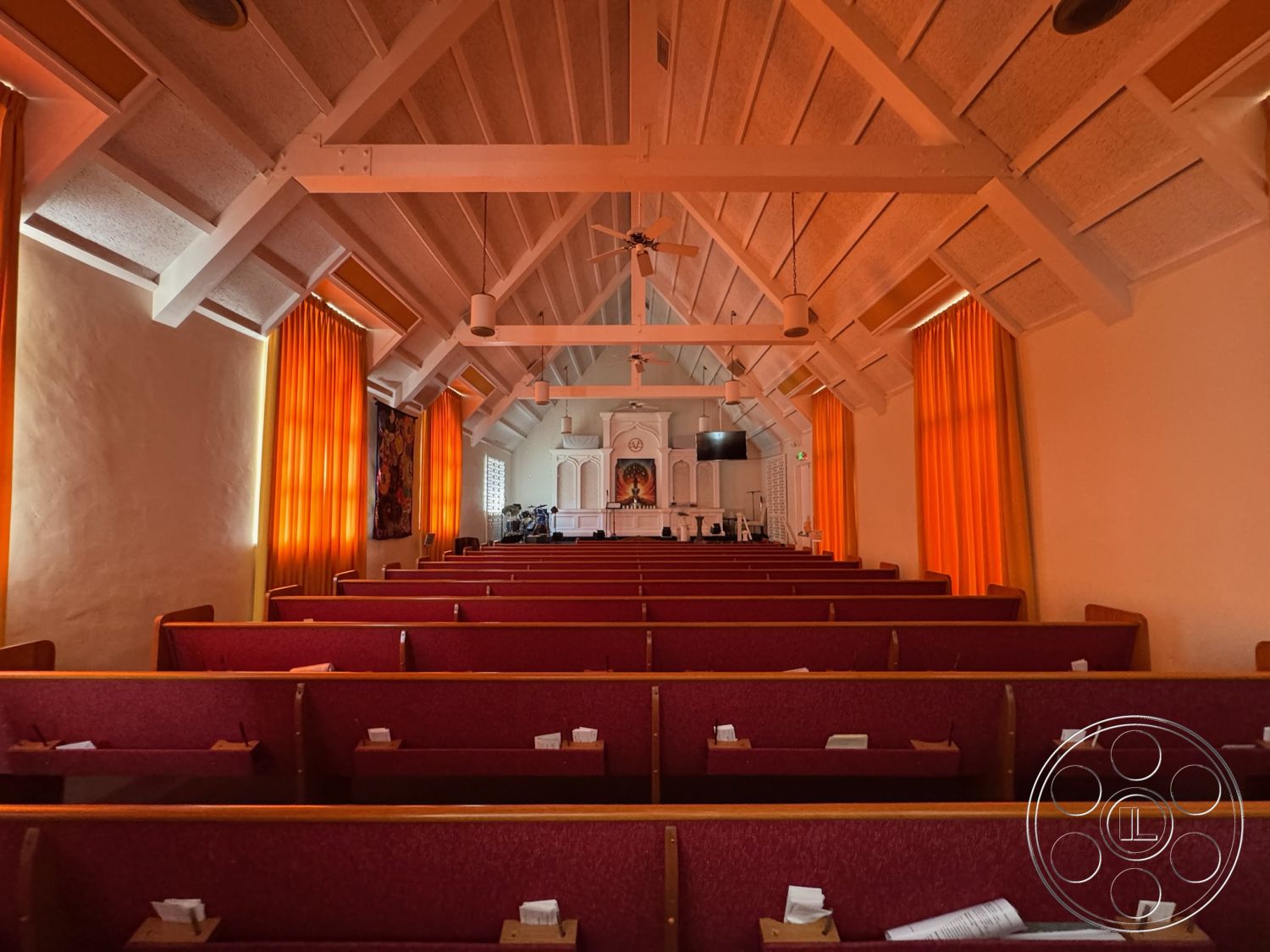 Spiritual Center - neutral color palette, natural light from windows, wood beam ceiling, white painted walls, interior church setting, exposed truss ceiling details, orange sheer curtain treatments, red upholstered church pews, wood paneling details, decorative altar area, flat screen TV installation, artistic wall hanging, architectural ceiling features, acoustic ceiling design, spacious gathering area