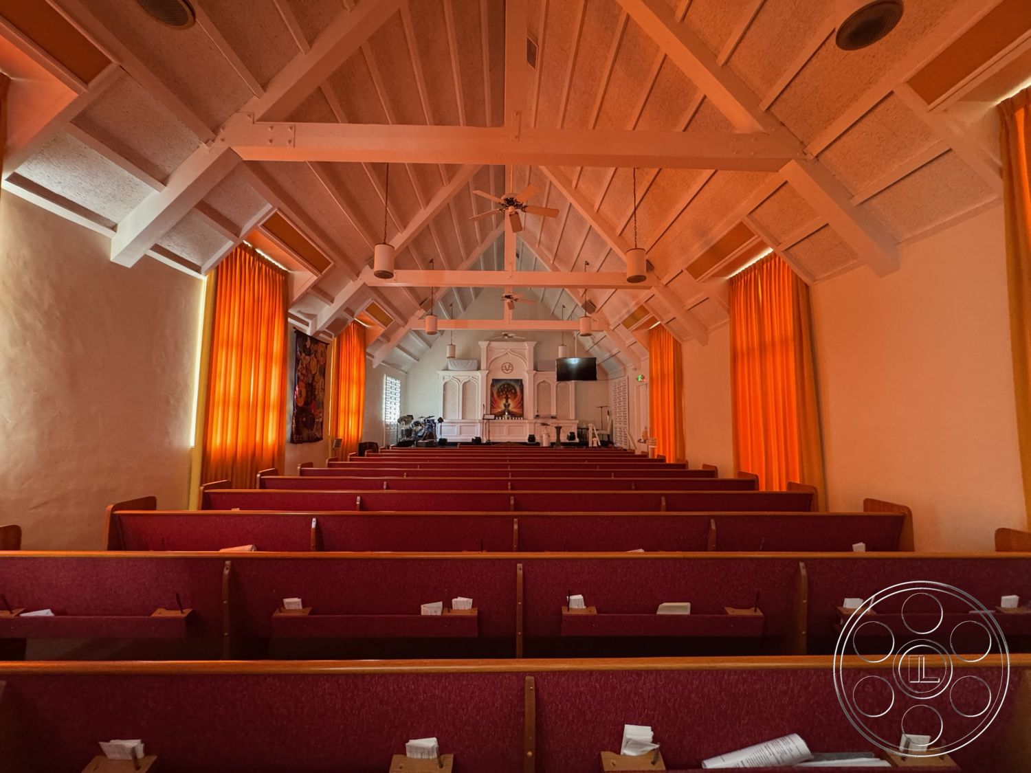 Spiritual Center - exposed wood ceiling beams interior, natural light from windows interior, orange fabric curtain wall interior, wooden pew seating interior, white plaster wall interior, raised platform stage interior, red upholstered pews interior, high vaulted ceiling interior, sound system equipment interior, decorative art wall interior, light wood trim details interior, cozy worship space interior